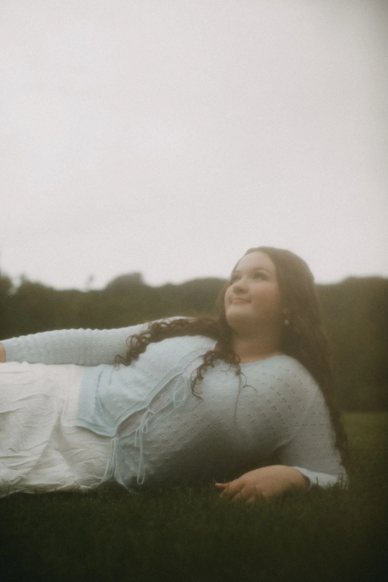 A woman with curly dark hair wearing a white dress with lace and drawstrings, lying on the grass outdoors on a cloudy day, smiling and looking into the distance.