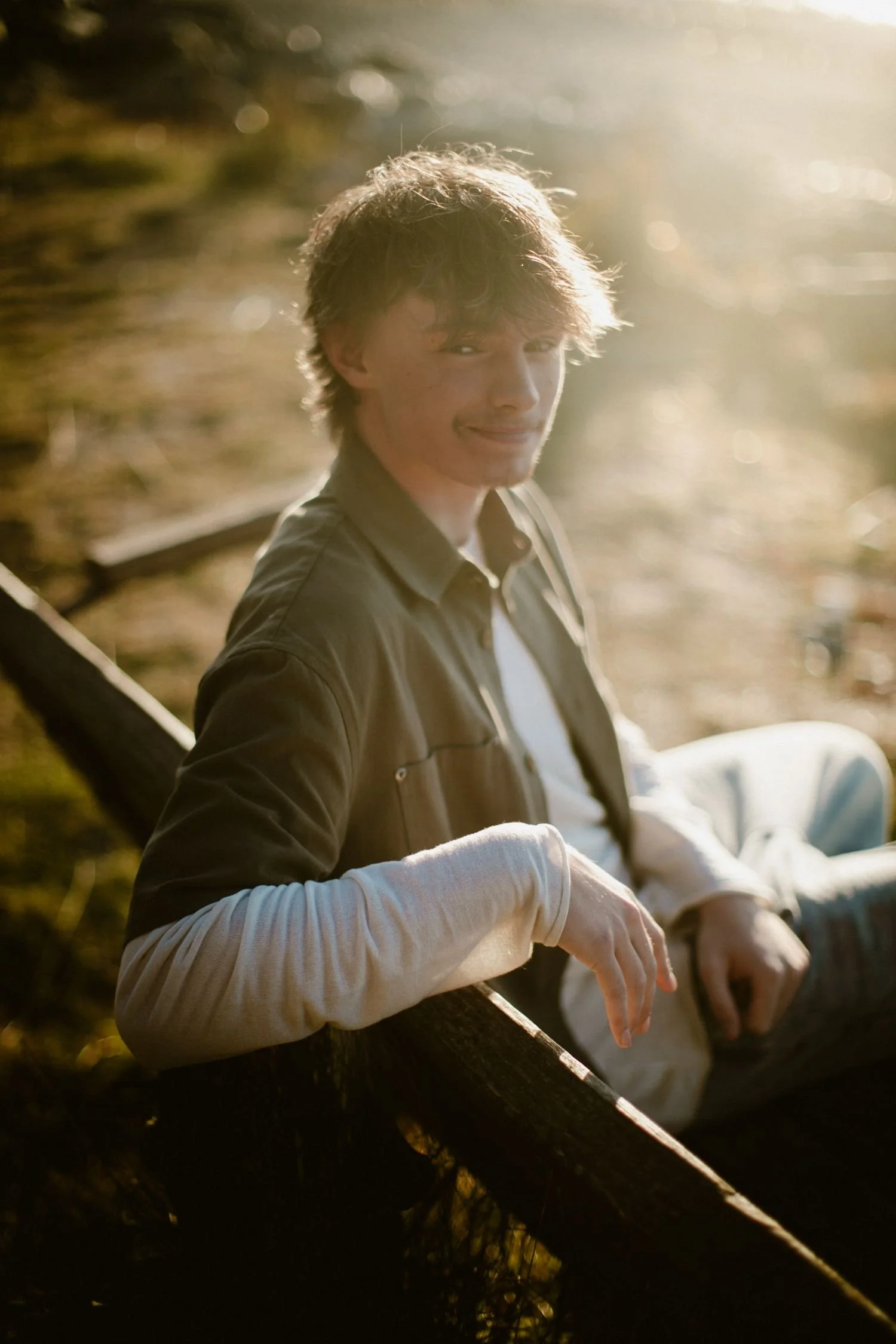 A young man with brown hair smiling, sitting outdoors on a fallen tree during sunset, with sunlight creating a warm glow and bokeh effect in the background.