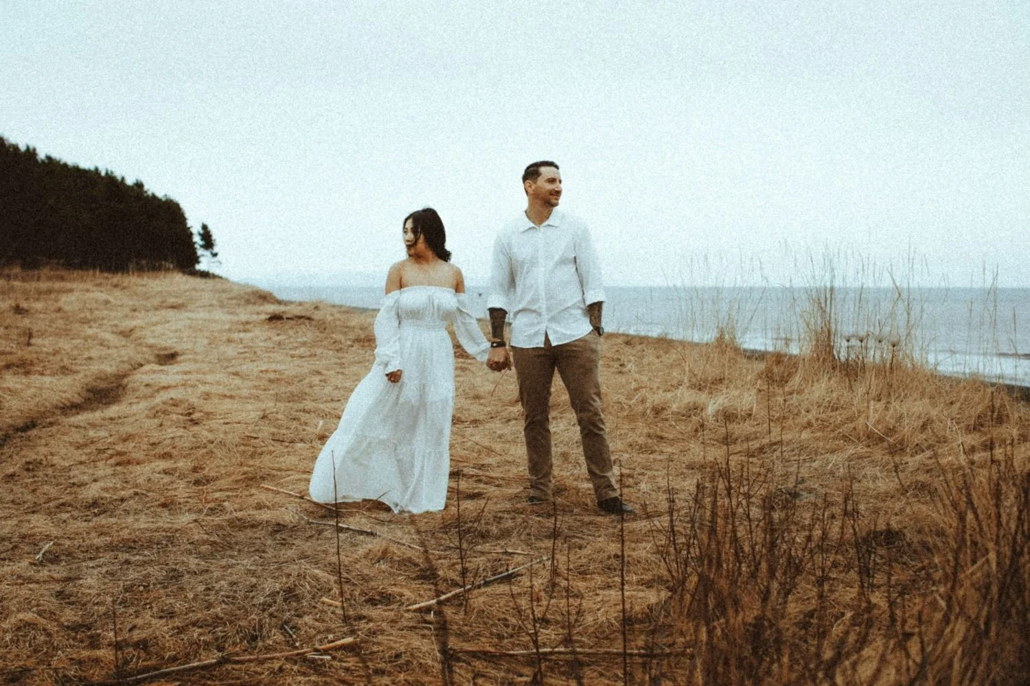 A couple holding hands walking along a sandy beach with dry grass and a body of water in the background, overcast sky.