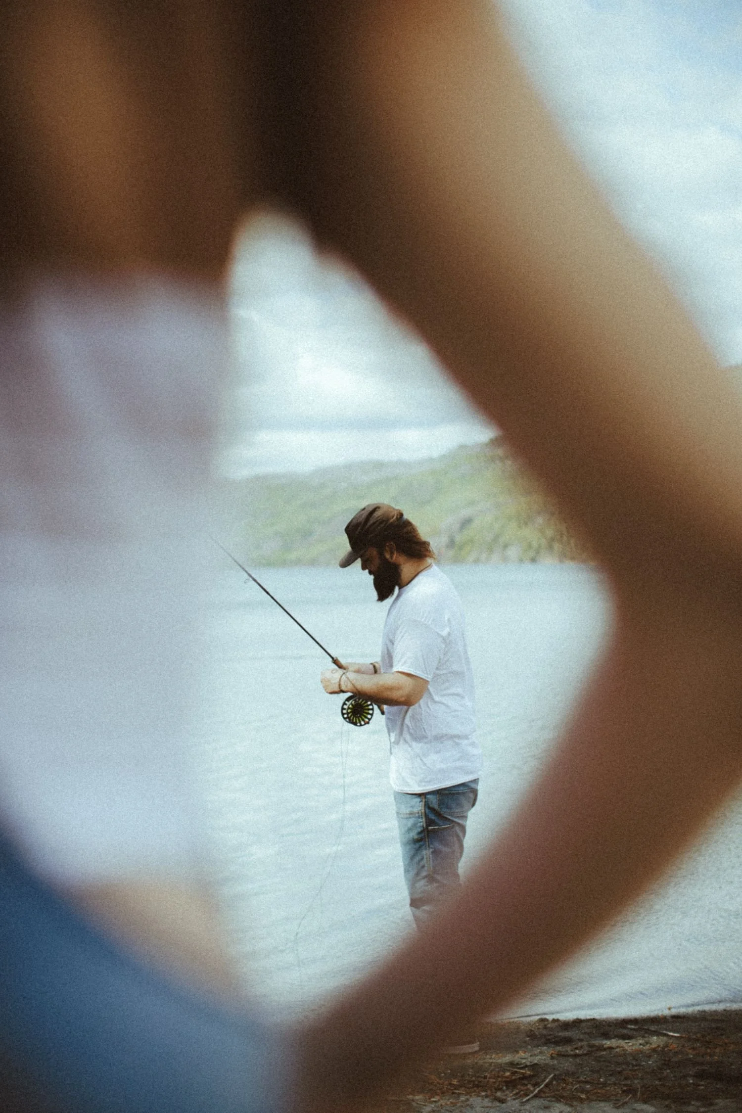 A man with long hair and a beard fishing in a body of water, wearing a gray cap, a white shirt, and blue jeans, seen through the blurry elbow of his partner.
