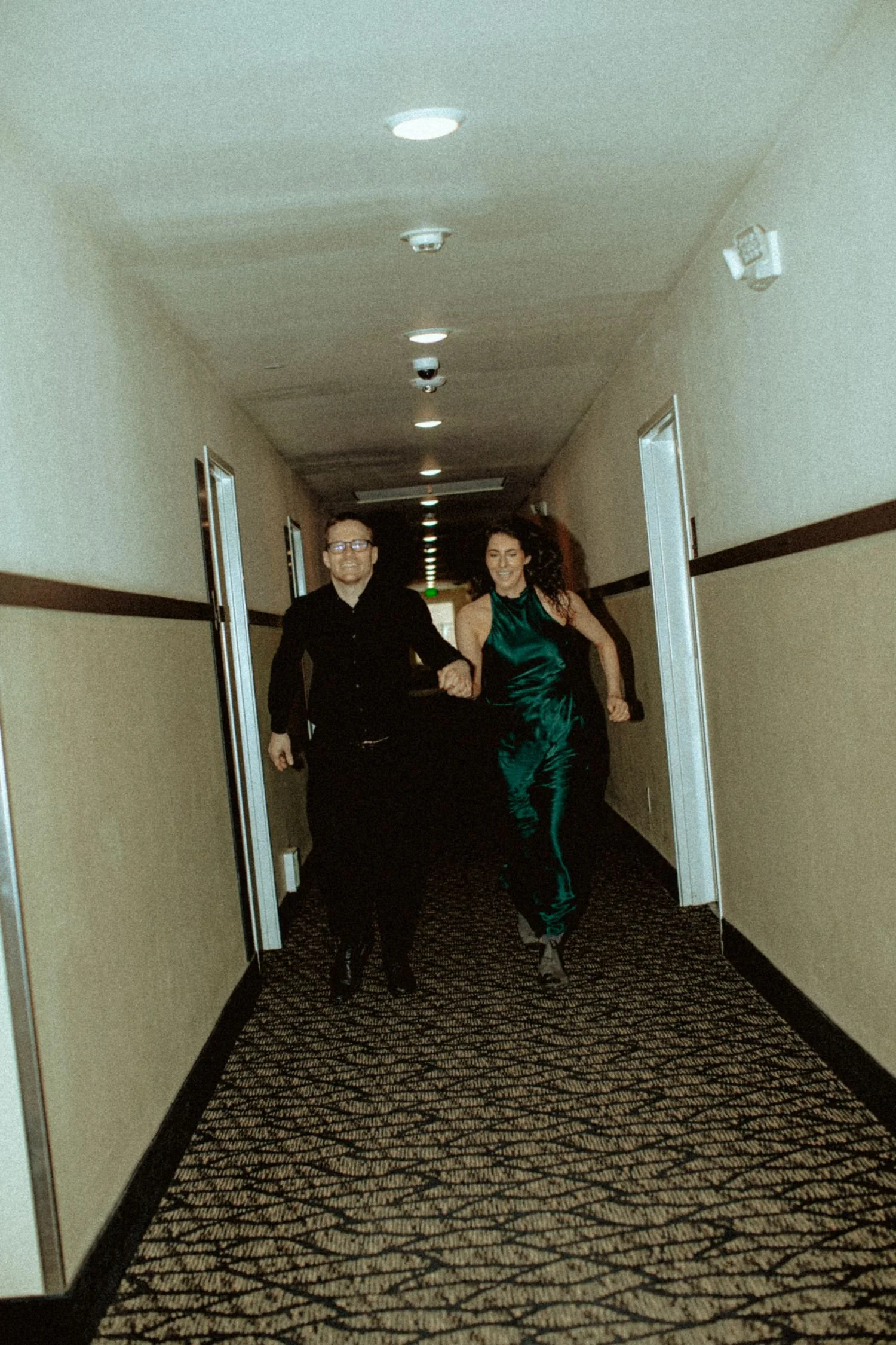 A man and a woman running down a hotel corridor, holding hands. The man is wearing a black shirt and glasses, and the woman is wearing a shiny green dress.