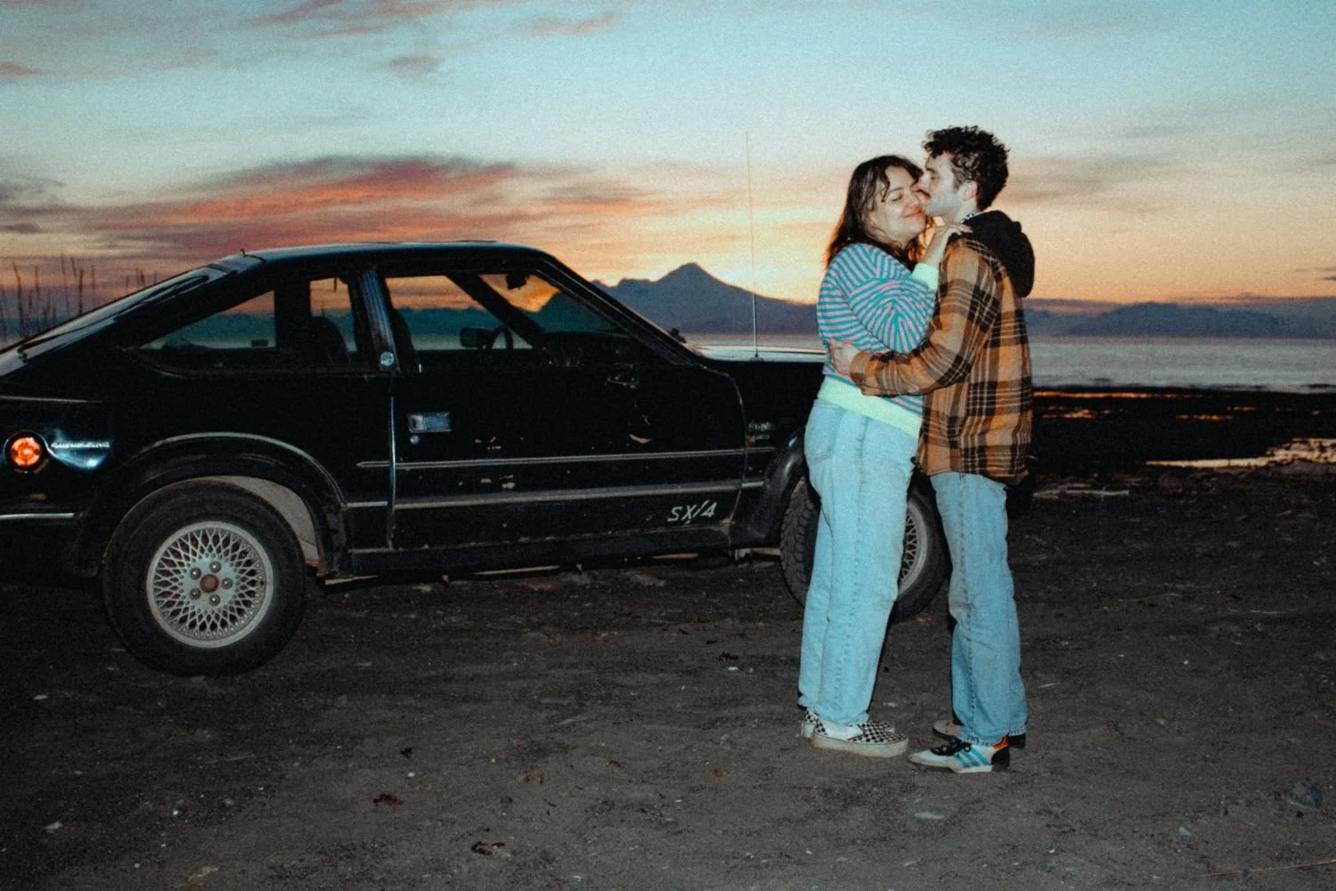 A young couple hugging and about to kiss next to a black vintage car on a beach at sunset, with mountains in the background.