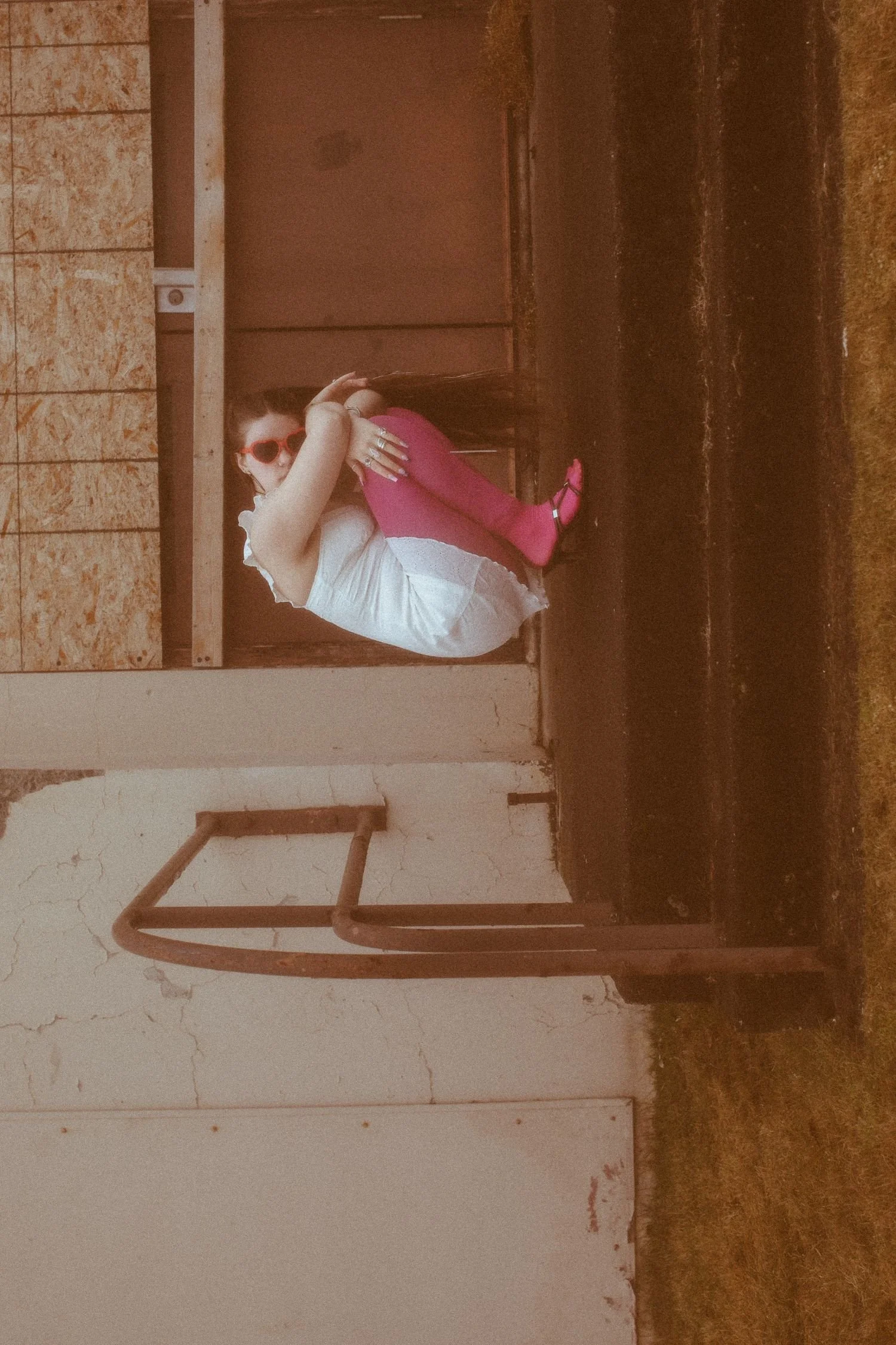A woman sitting on a step, wearing pink tights, black sandals, a white blouse, and red sunglasses, hugging her knees with her hands, in an unfinished or abandoned building with exposed wallboards and peeling paint.