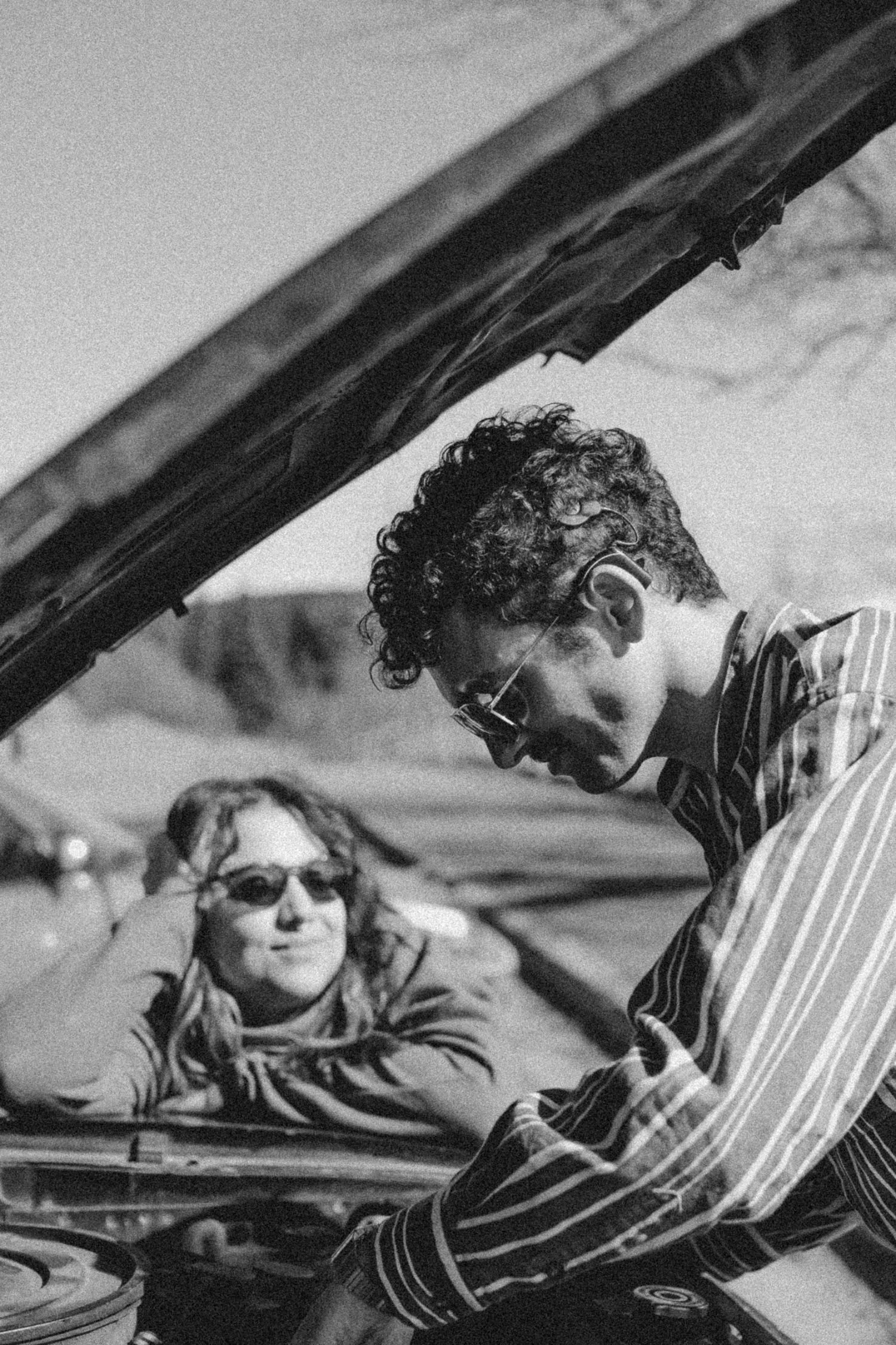 A young man with curly hair wearing glasses and a striped shirt playing a piano, with a woman in sunglasses resting her head on her hand in the background.