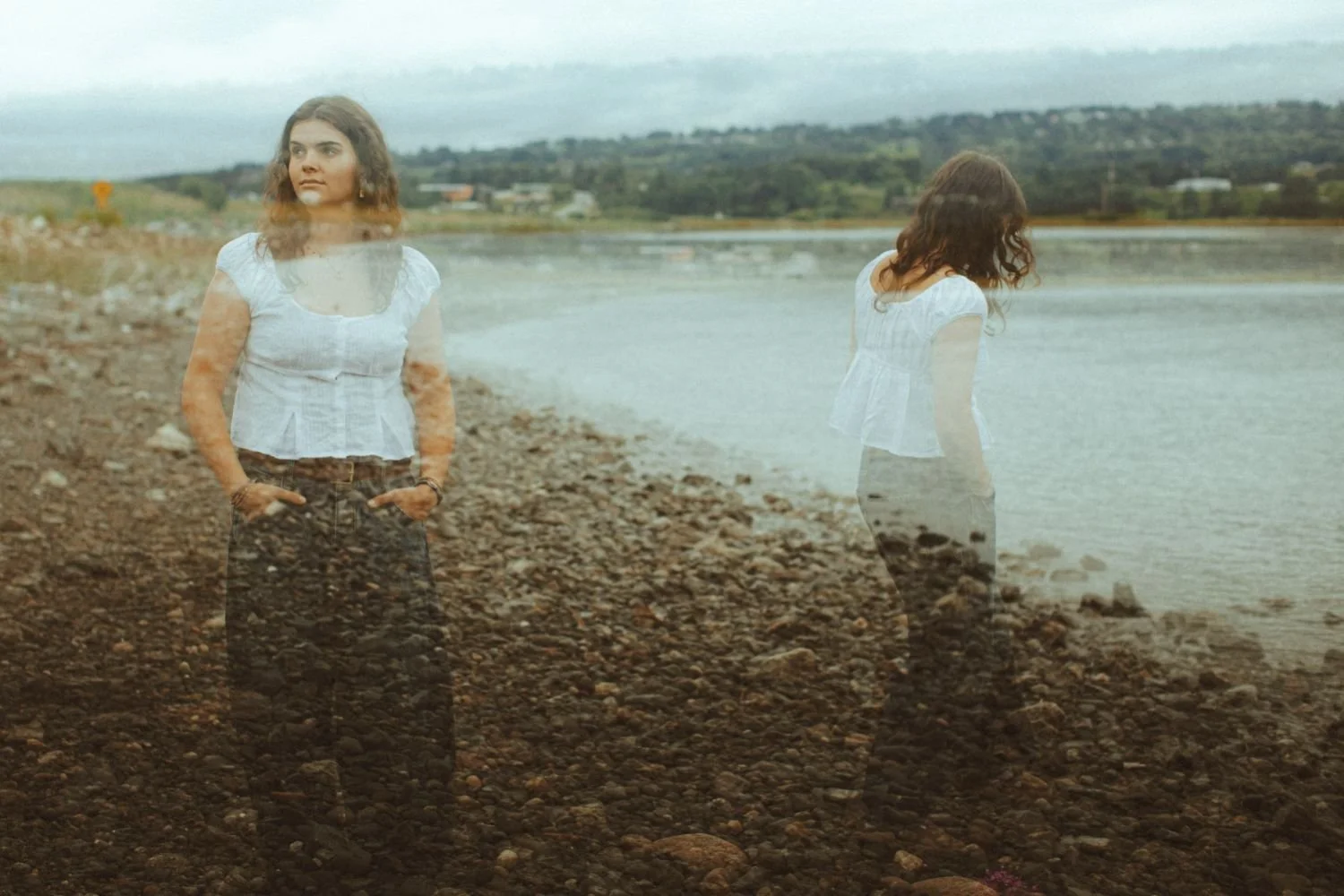 A double exposure photograph of a woman standing on a rocky shoreline, with her reflection appearing as a ghostly image overlapping her body. The scene includes the water, distant land, and cloudy sky.