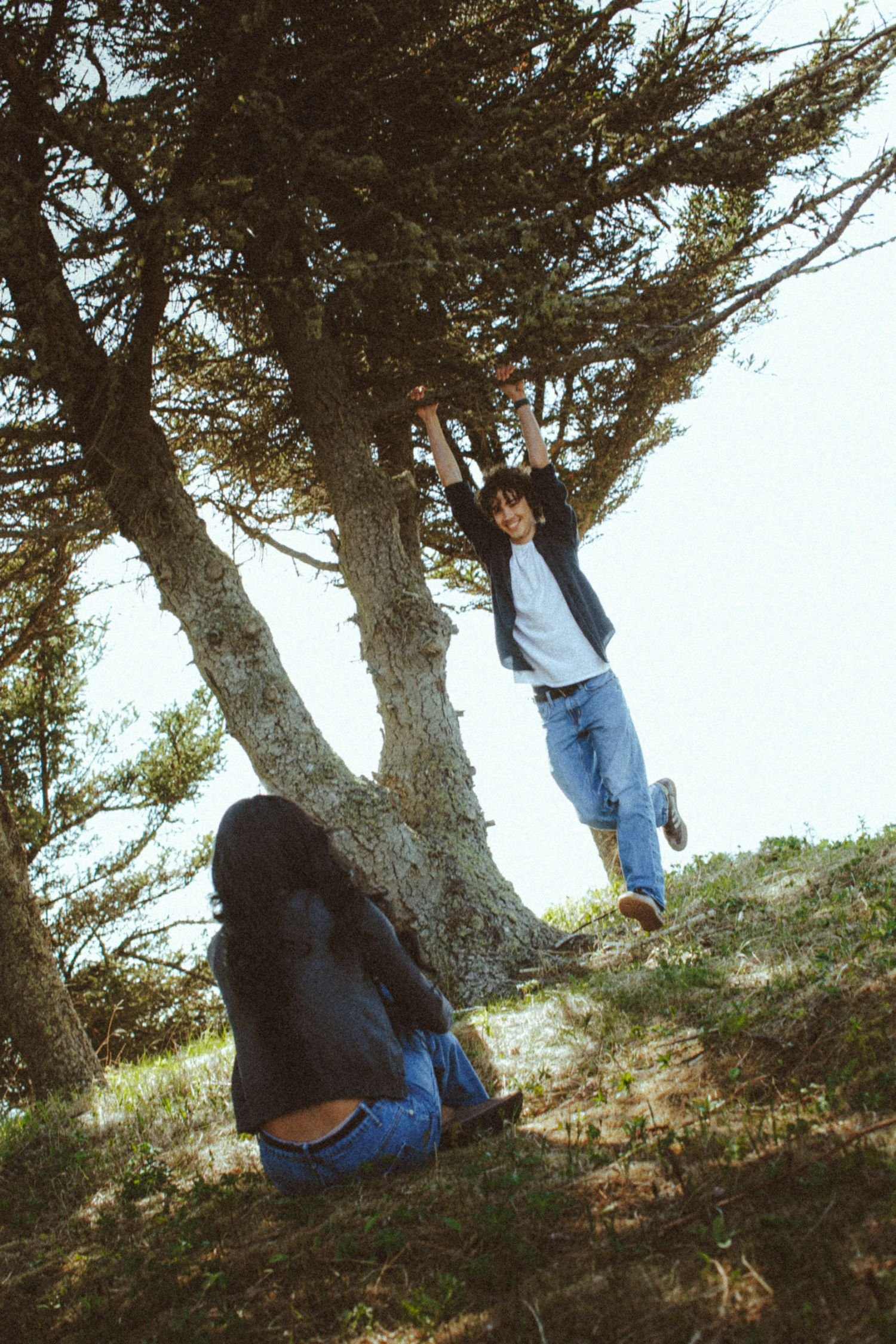 A young man in a dark jacket and blue jeans is hanging from a large tree branch, smiling. A girl in a black jacket and blue jeans is sitting on the ground beneath the tree, looking up at him.