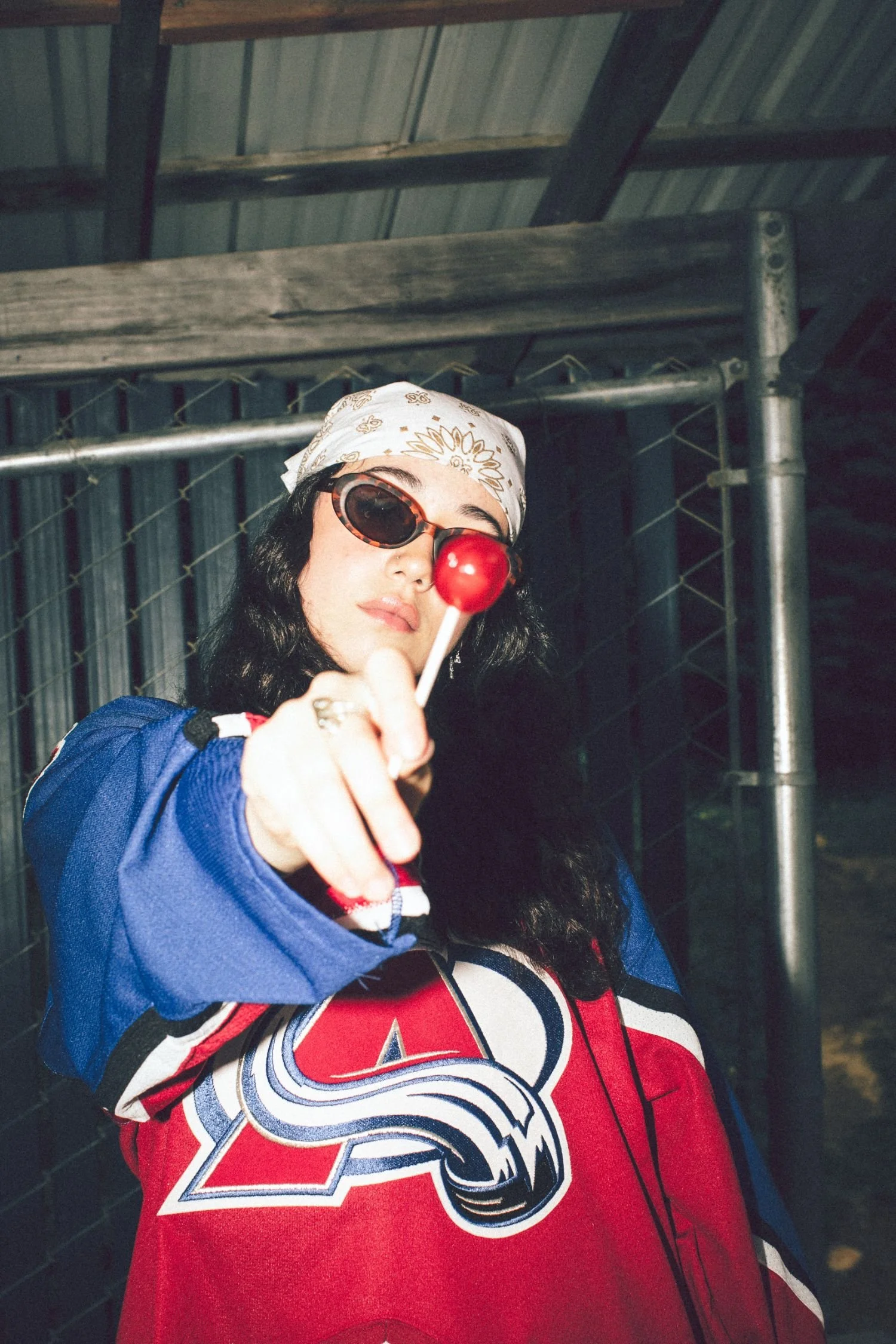 A person with long black hair wearing sunglasses, a white bandana, and a red and blue sports jersey, pointing towards the camera in a dark industrial setting.