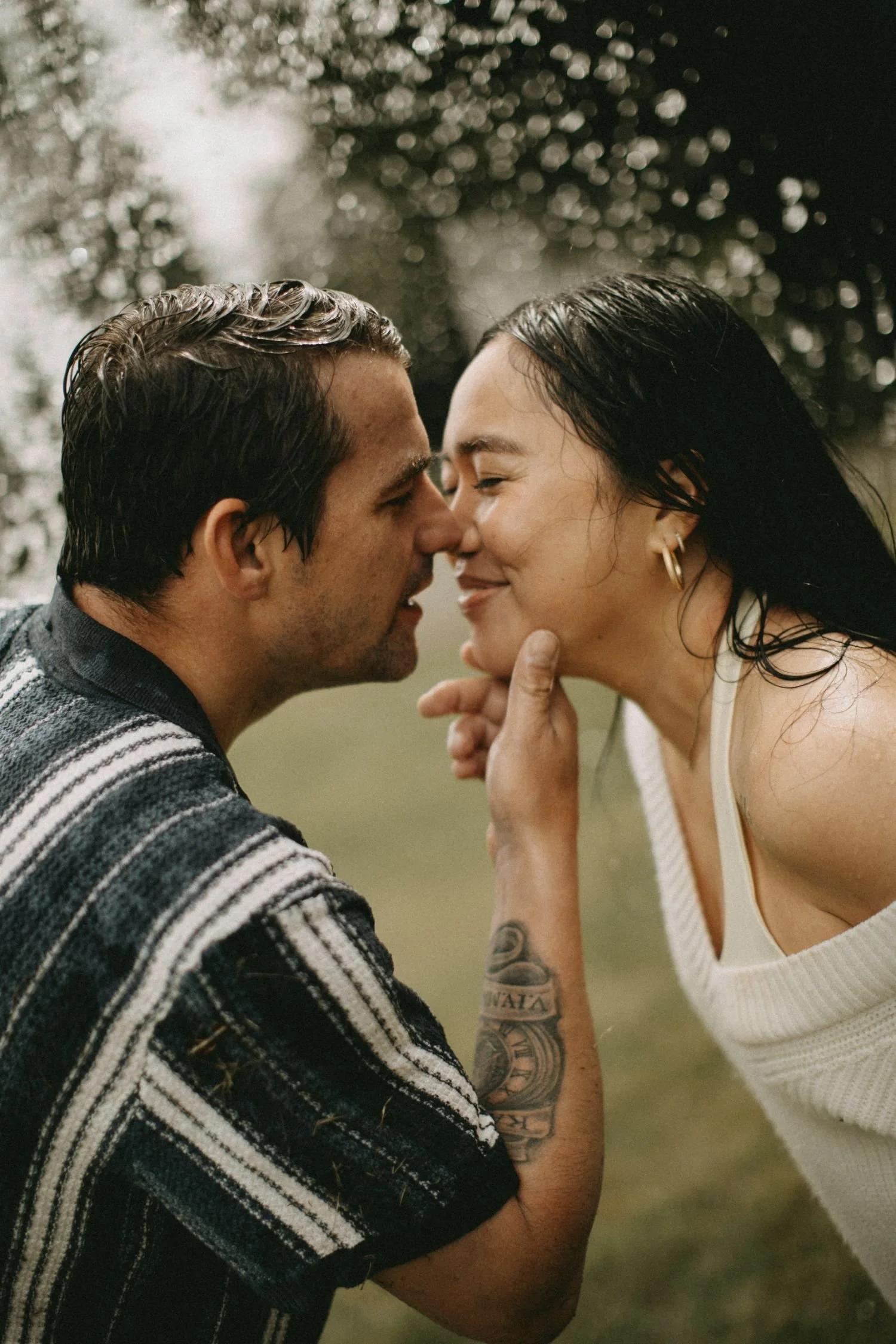 A couple touching foreheads and smiling in a close-up photo, outdoors with blurred trees in the background.