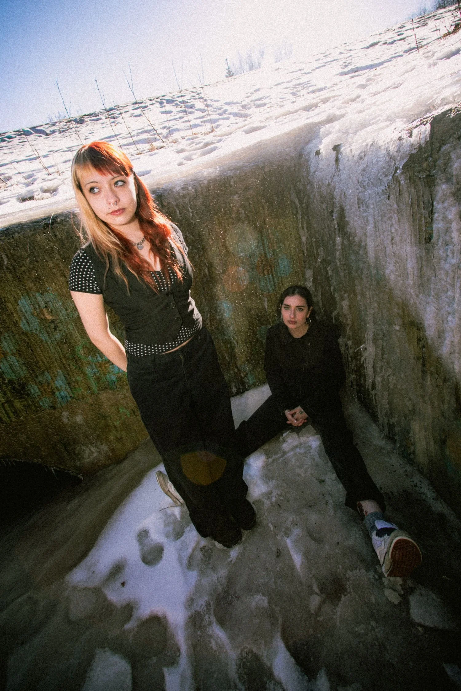 Two young women in a small, snow-covered cave or tunnel. One standing, the other sitting with snow on the ground, surrounded by a snowy landscape with leafless trees and a fence in the background.