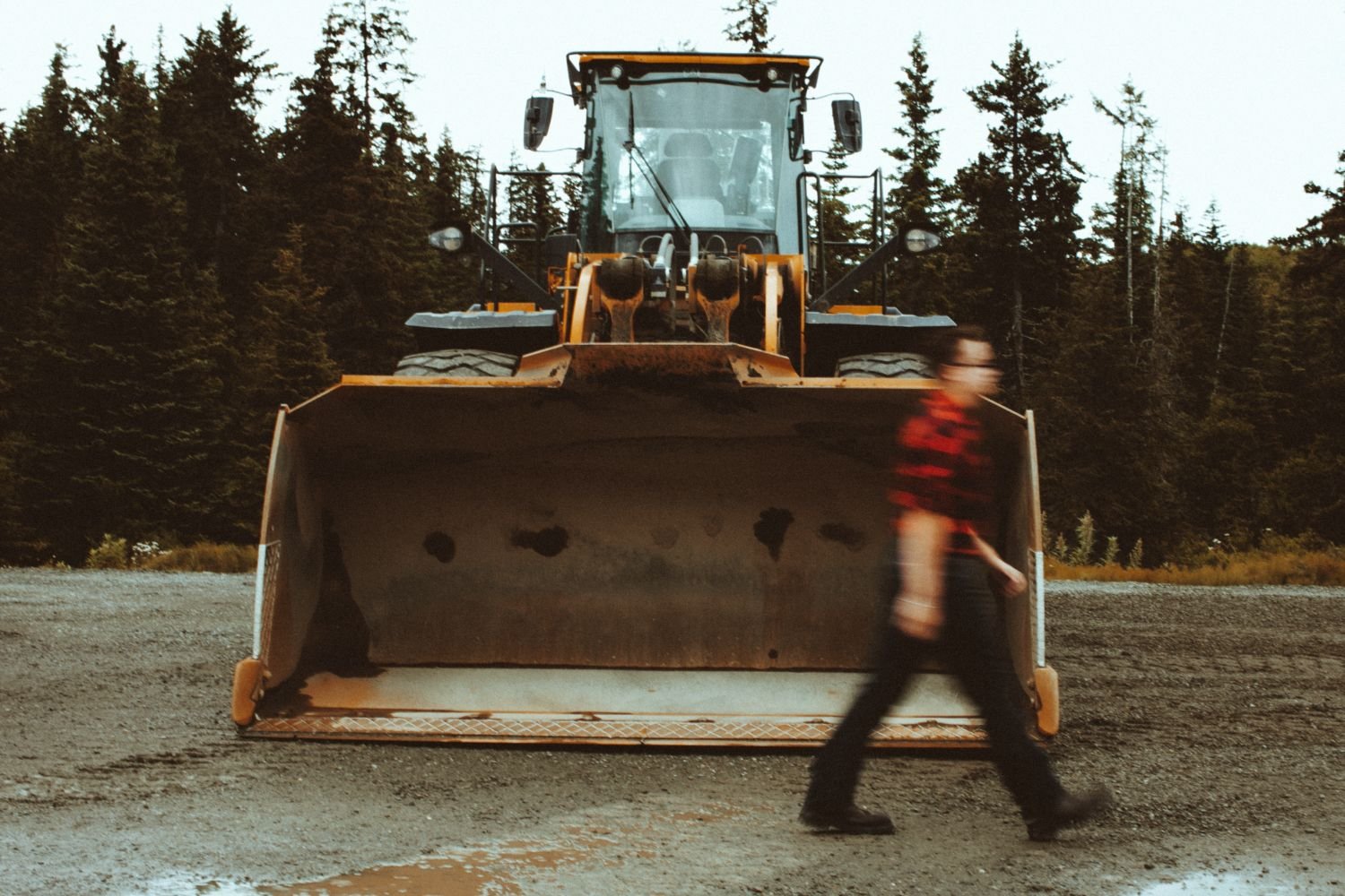 A person walking past a large yellow bulldozer on a gravel surface with a background of trees.