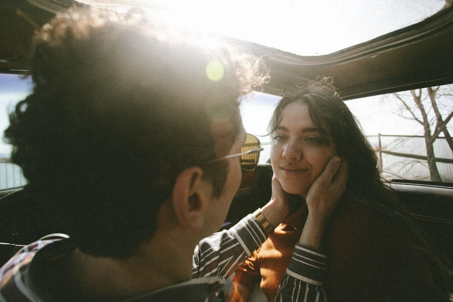 A man and a woman in a car, with the woman touching the man's face and smiling at him during daylight.