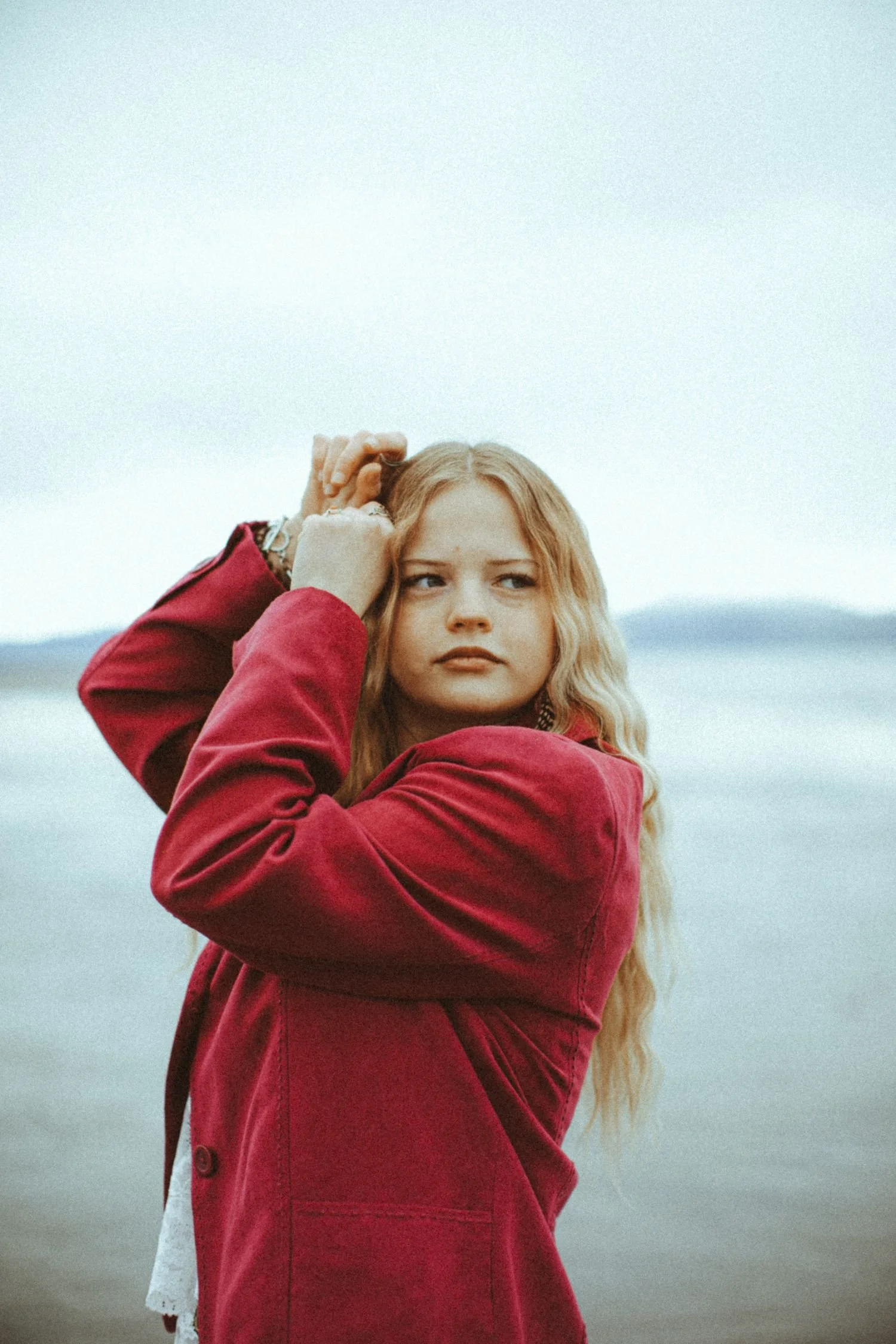 A young woman with long blonde hair wearing a red jacket, standing outdoors against a pale sky and distant landscape background.