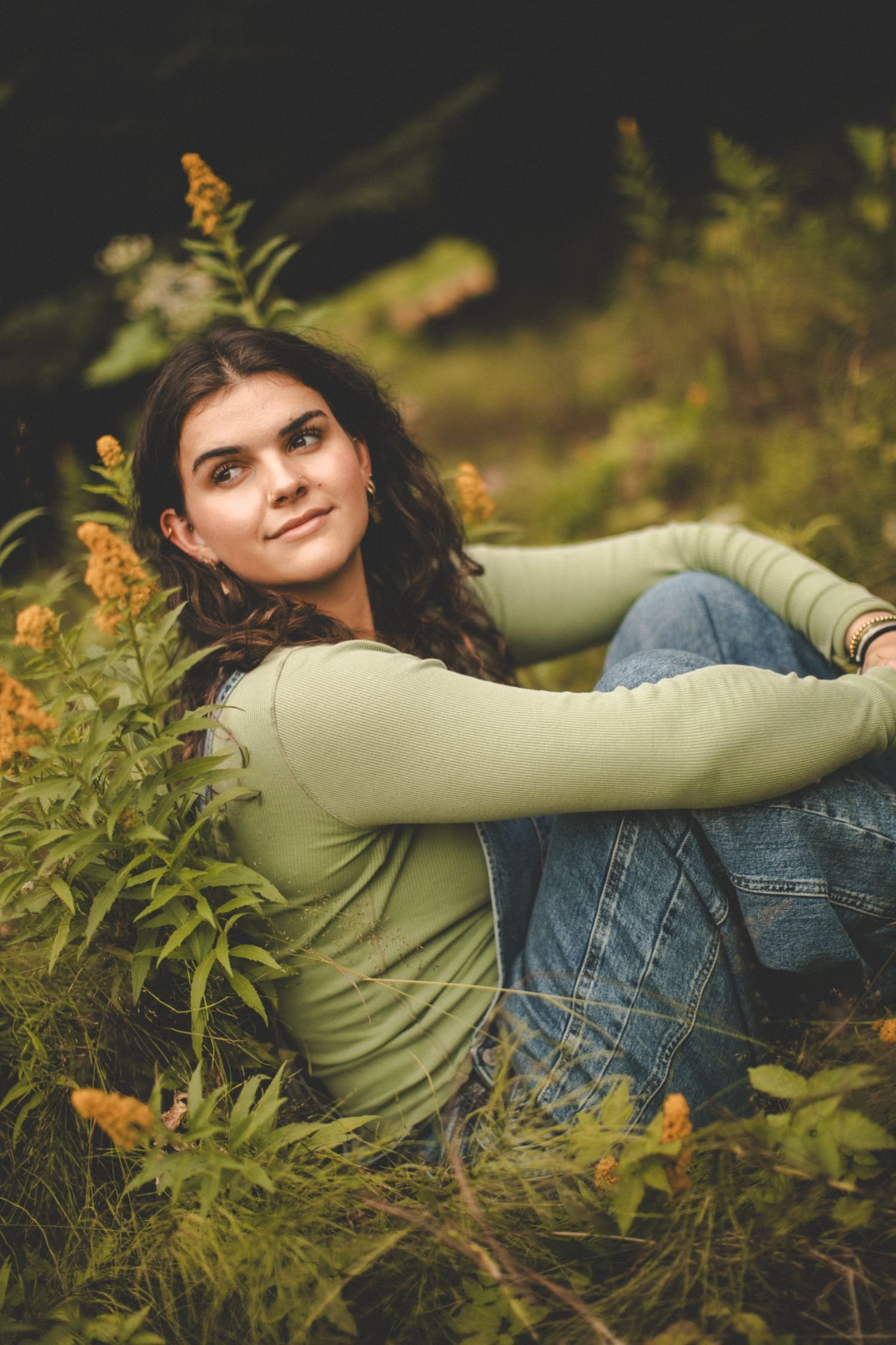 A young woman with dark, wavy hair and a slight smile, lying in a field of green plants and yellow flowers, wearing a light green long-sleeve shirt and blue jeans, with a blurred background of greenery.