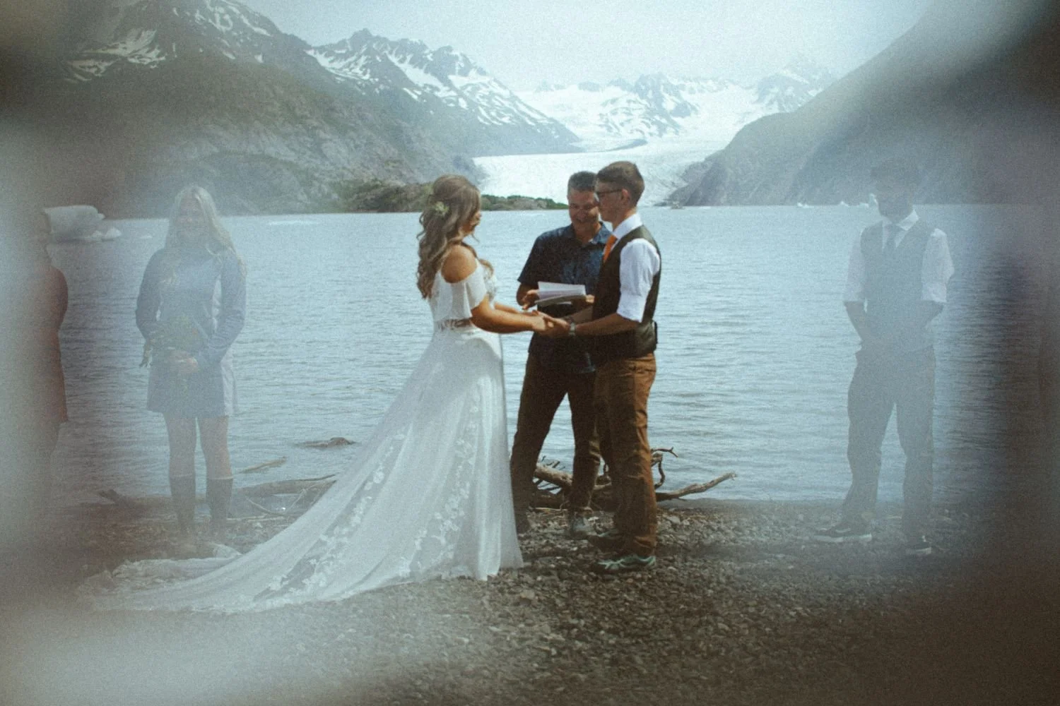 A wedding ceremony takes place by a lake with snowy mountains in the background, featuring a bride and groom exchanging vows, with three people standing nearby.