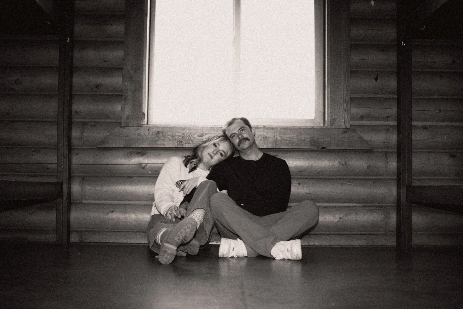 Black and white photo of a man and woman sitting on the floor, leaning against a wooden wall with a large window above them. The woman has her head resting on the man's shoulder, and both are looking slightly upward.