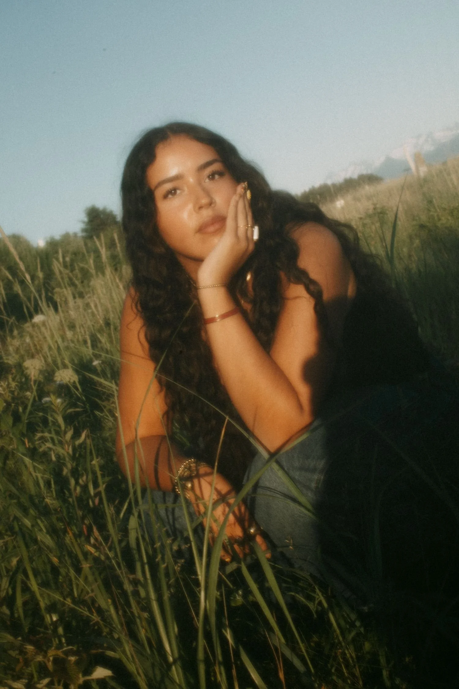A young woman with long dark curly hair sitting outdoors in a grassy field during sunset, resting her face on her hand, wearing multiple rings and bracelets, looking at the camera.