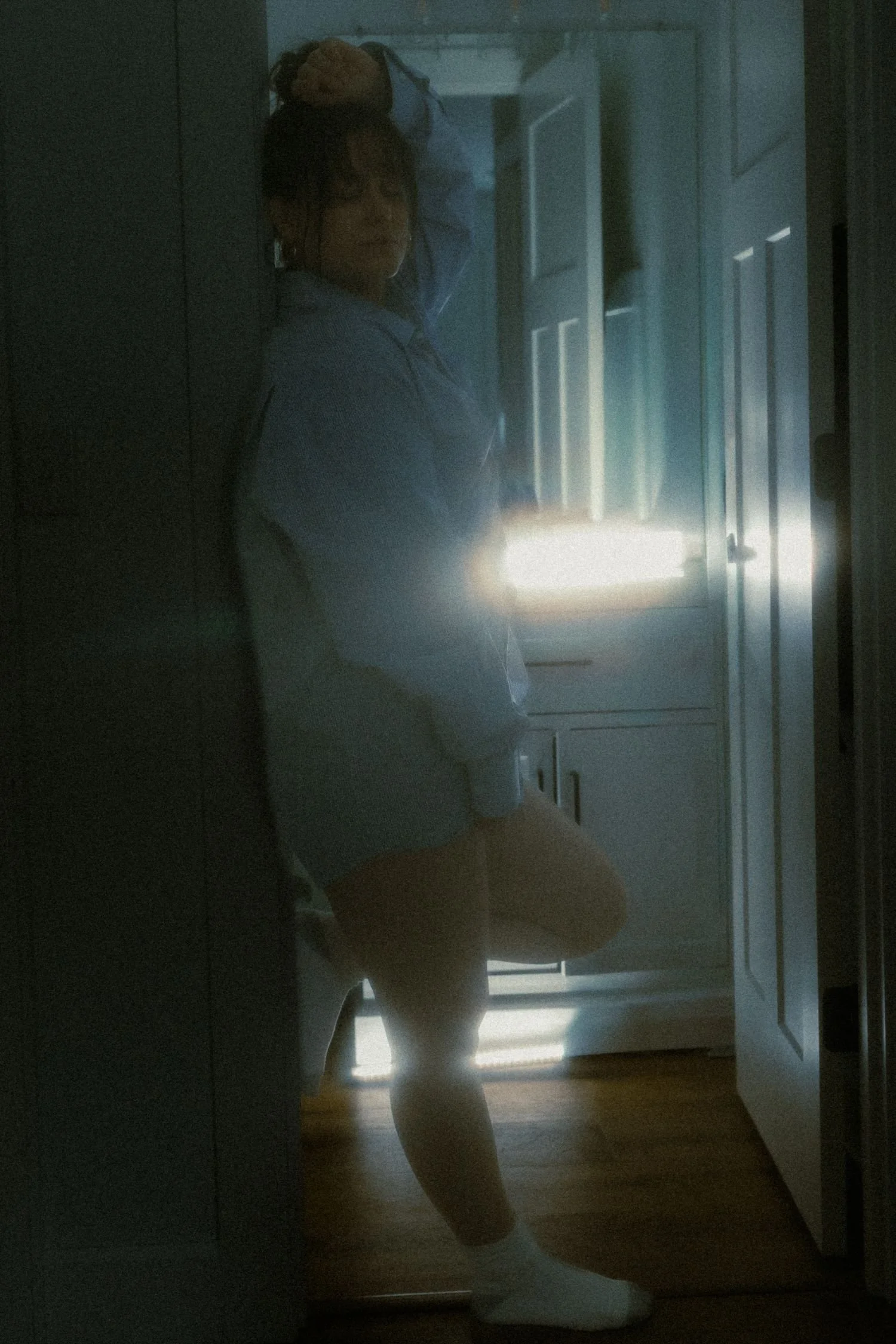 A woman in pajamas standing in front of a mirror, with natural light coming through a window behind her.