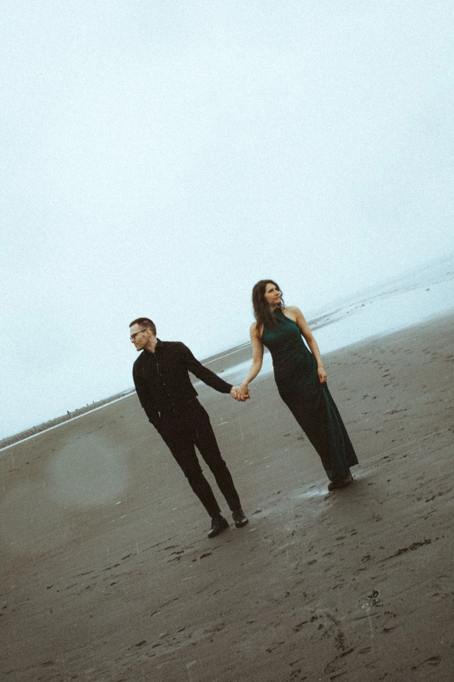 A man and woman holding hands on a beach with overcast sky, the woman in a dark green dress and the man in black.