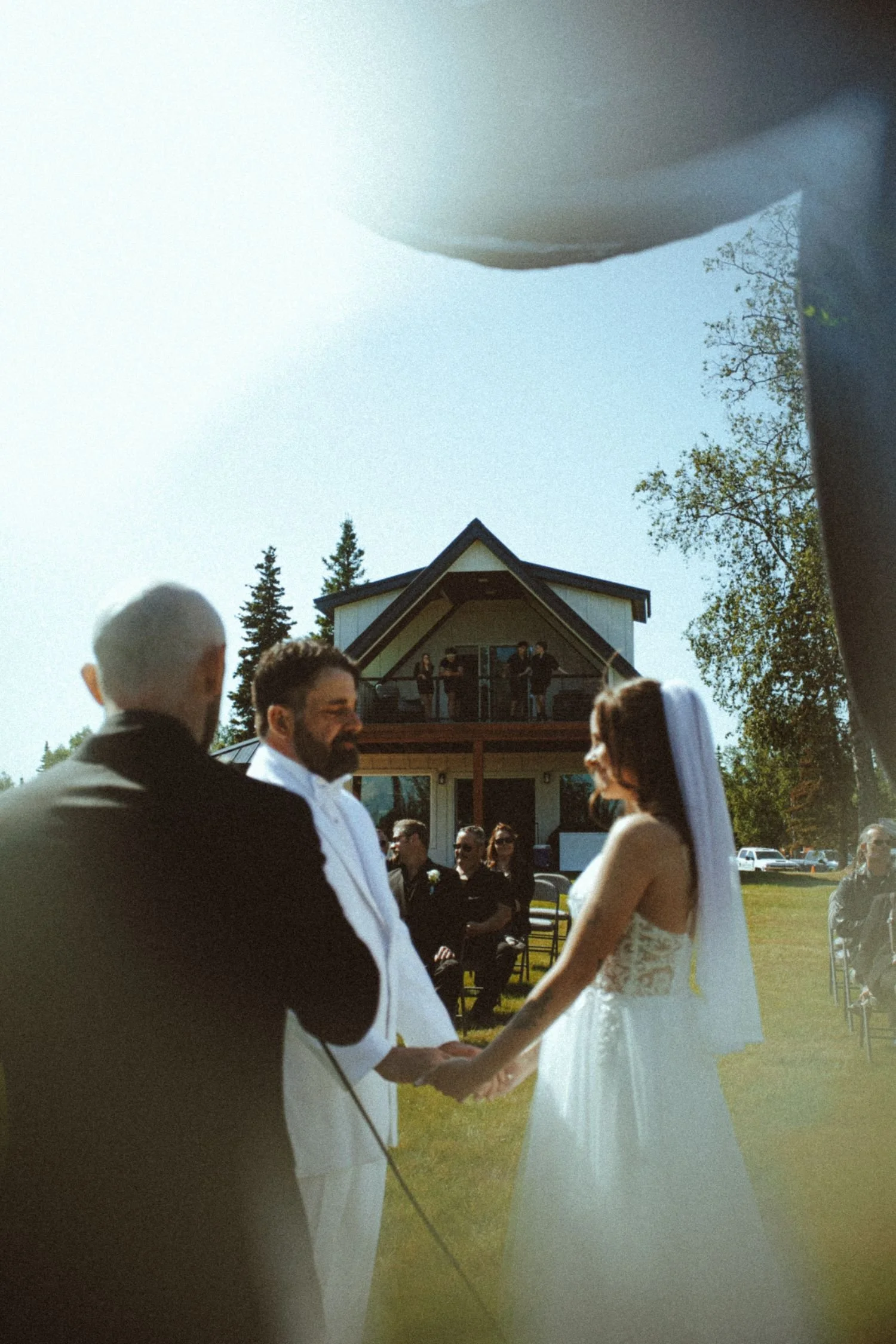 A bride and groom standing outdoors during their wedding ceremony, holding hands and facing each other, surrounded by guests, with a house and trees in the background.