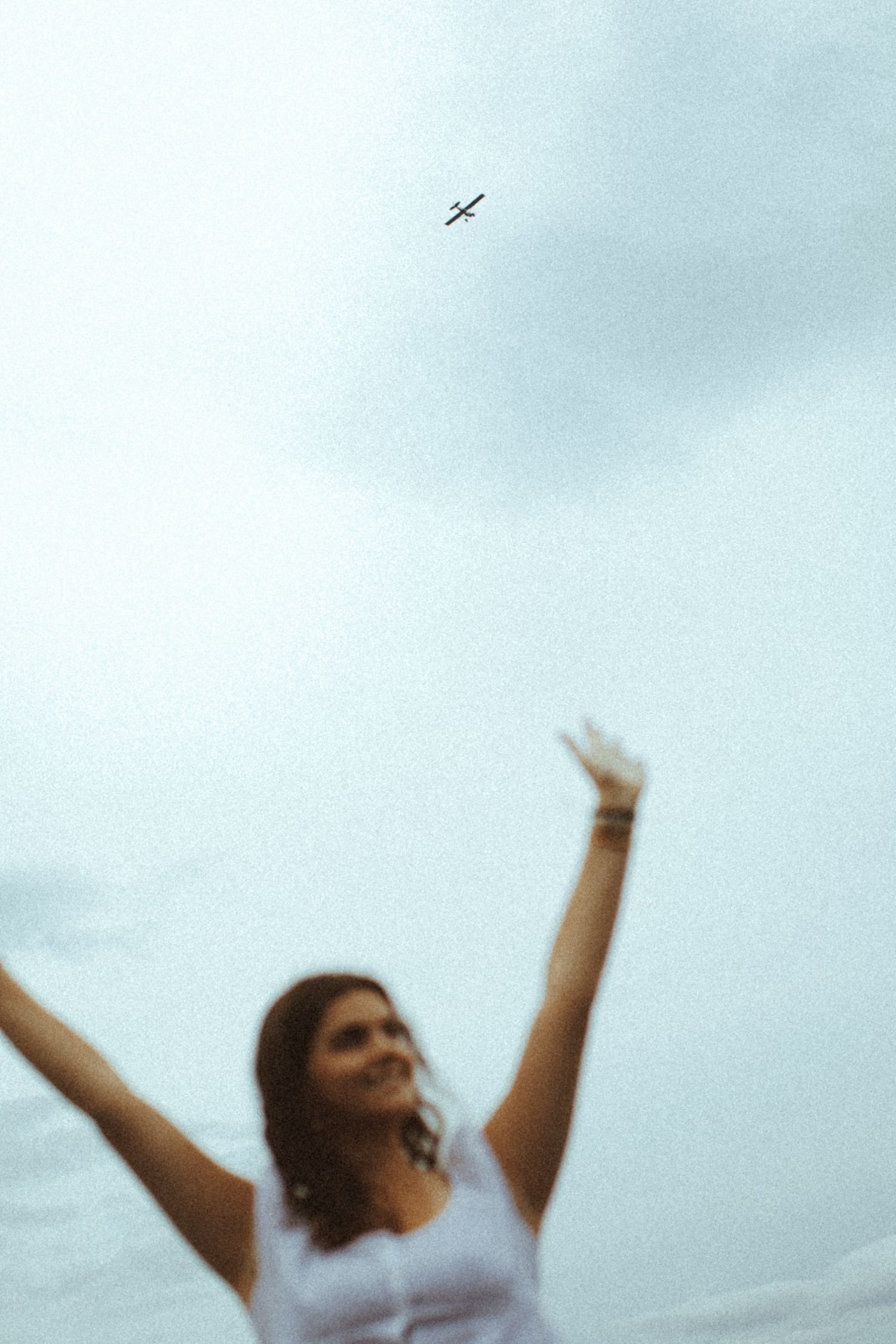 A woman with long hair reaching upward, smiling and raising her arms in a joyful gesture against a cloudy sky, with a small airplane flying in the background.