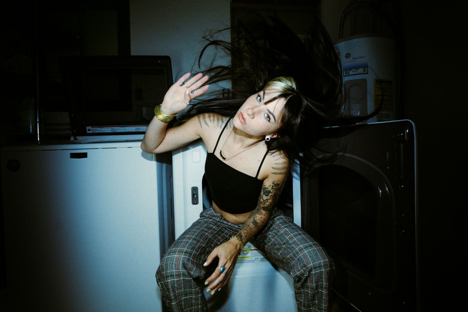 Danika Styles with long dark hair, tattoos, and a black top sitting on a washing machine in a dimly lit laundry room, looking at the camera.