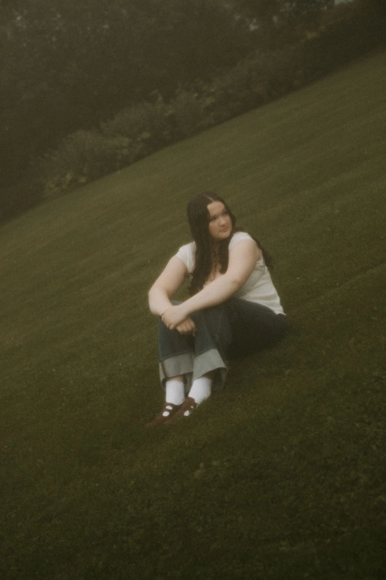 A young woman with long dark hair, wearing a white top and dark pants with white knee-high socks, sitting on the grass under a large tree at night.