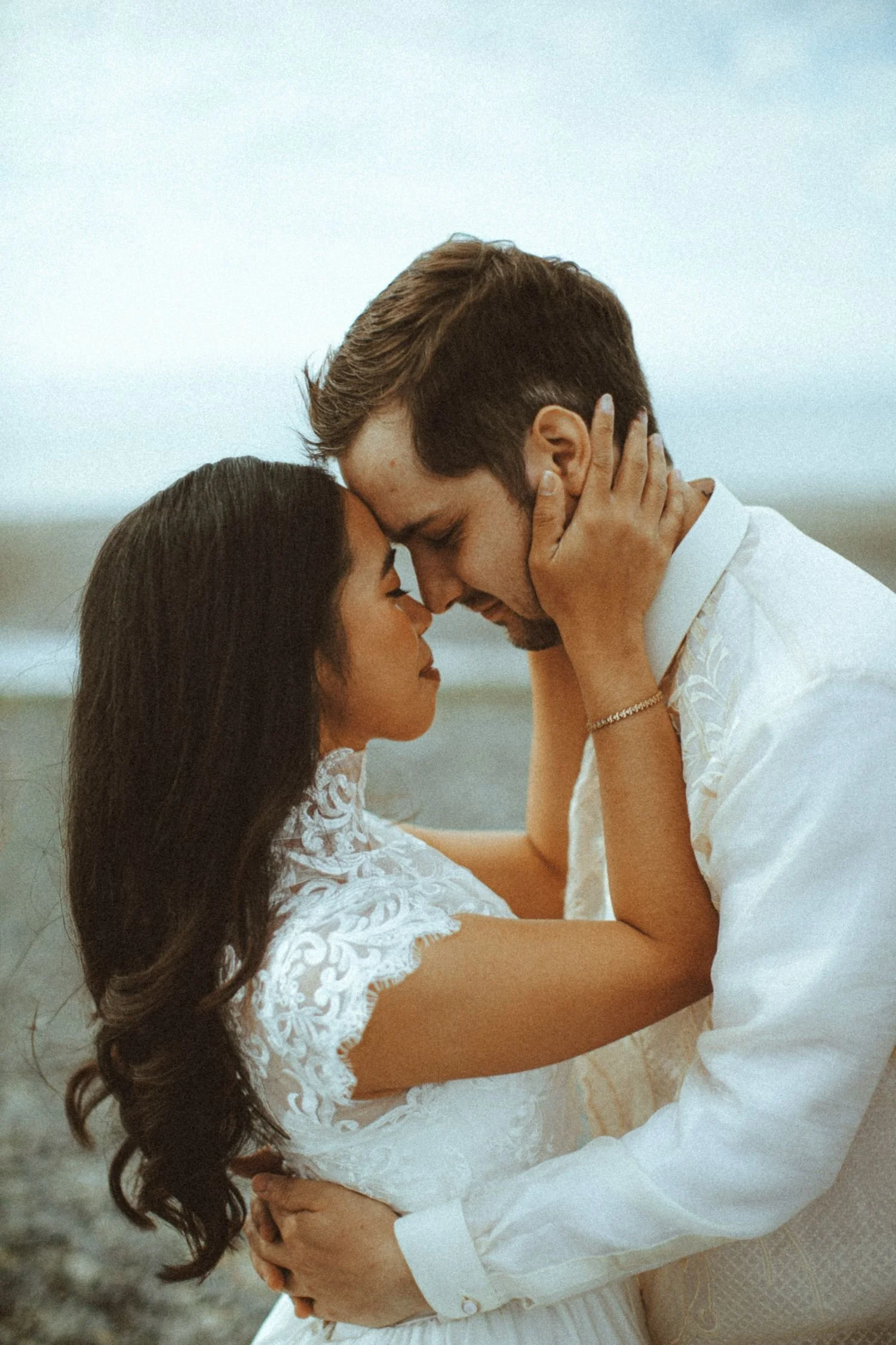 A couple in wedding attire sharing an intimate moment on the beach, with the woman gently touching the man's face. The woman has long dark hair and wears a white lace dress. The man has short brown hair and a beard, wearing a white shirt. They are standing close, with their foreheads touching and eyes closed, on a cloudy day near the ocean.