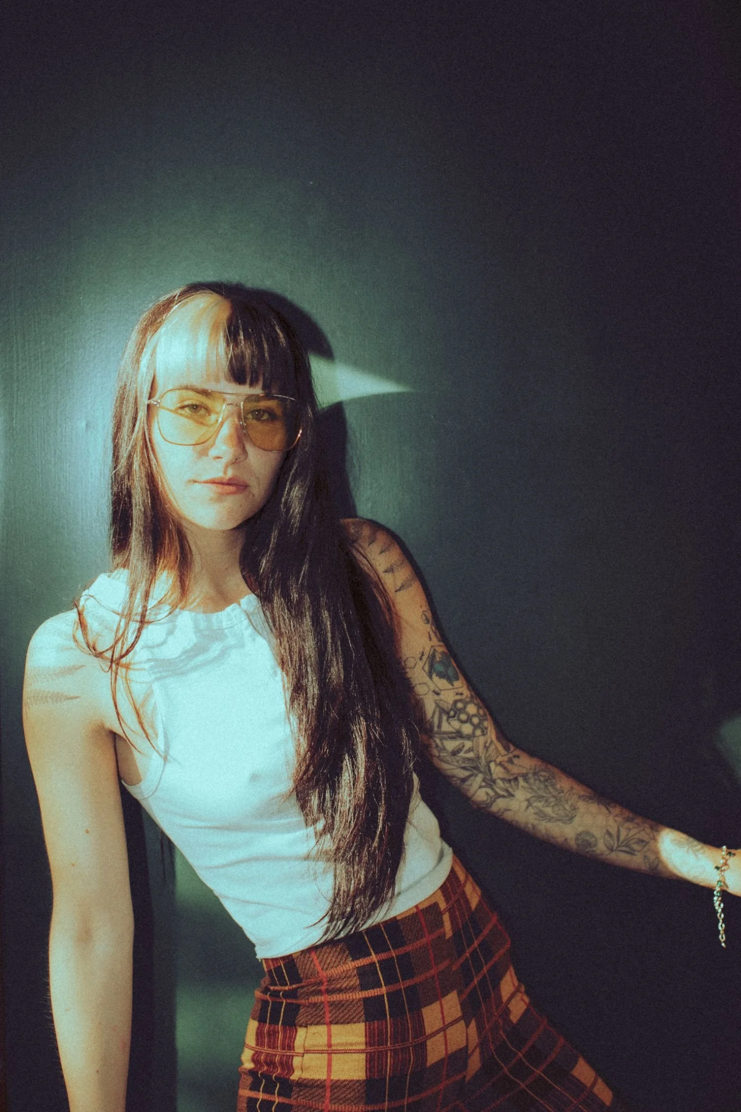 Danika Stiles with long dark hair that has blonde bangs, wearing large yellow-tinted glasses and a sleeveless white top. She has tattoos on her right arm and is standing against a dark wall, with confident expression and a slight smile.