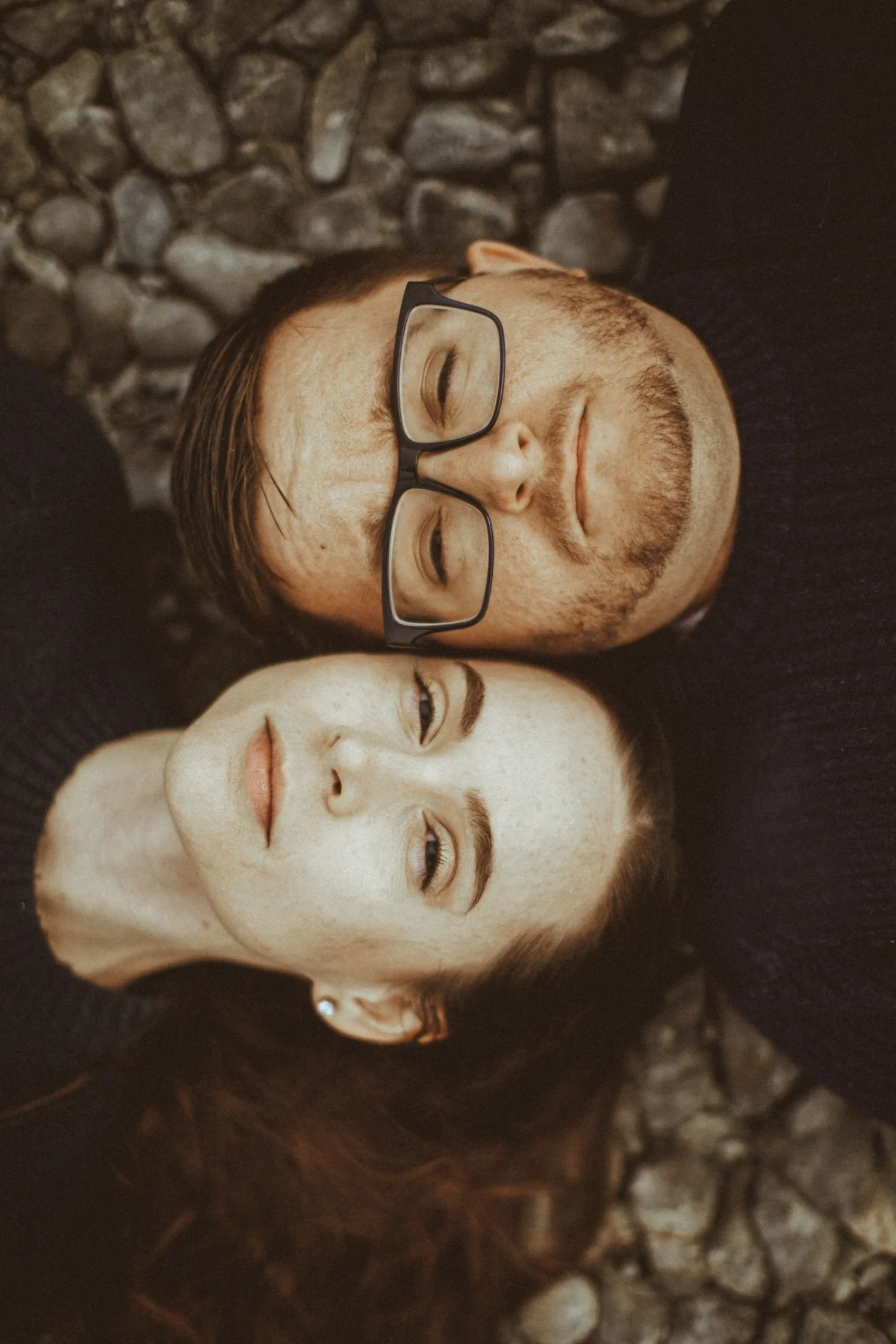 A man with glasses and a beard lying on a bed with a woman with long dark hair and pierced ears, both with their eyes closed.