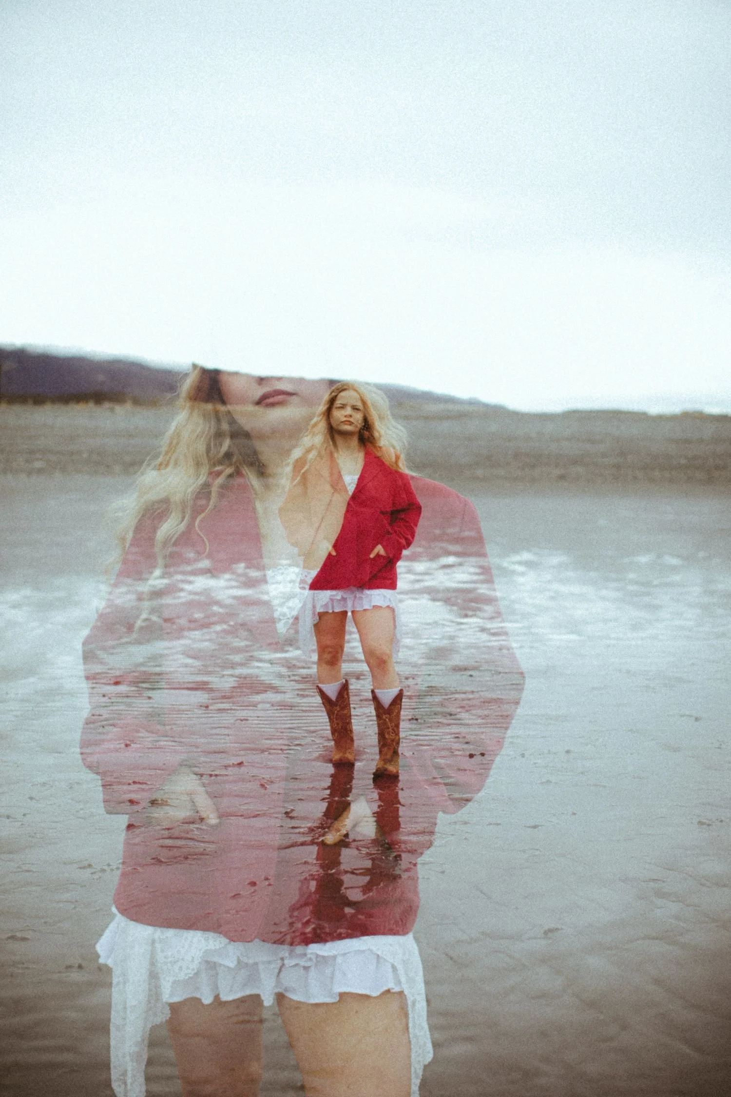 A person standing on a beach wearing cowboy boots, a red jacket, and a white dress with lace details, with a double exposure of a woman's face over the scene.