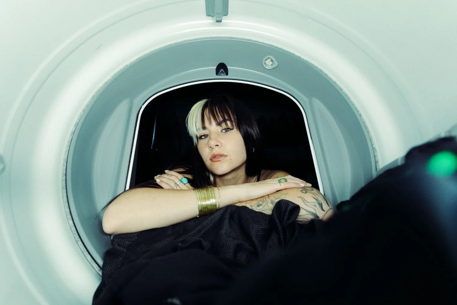 Danika Stiles with black and white hair leaning her head on her crossed arms inside a washing machine or dryer, looking at the camera.