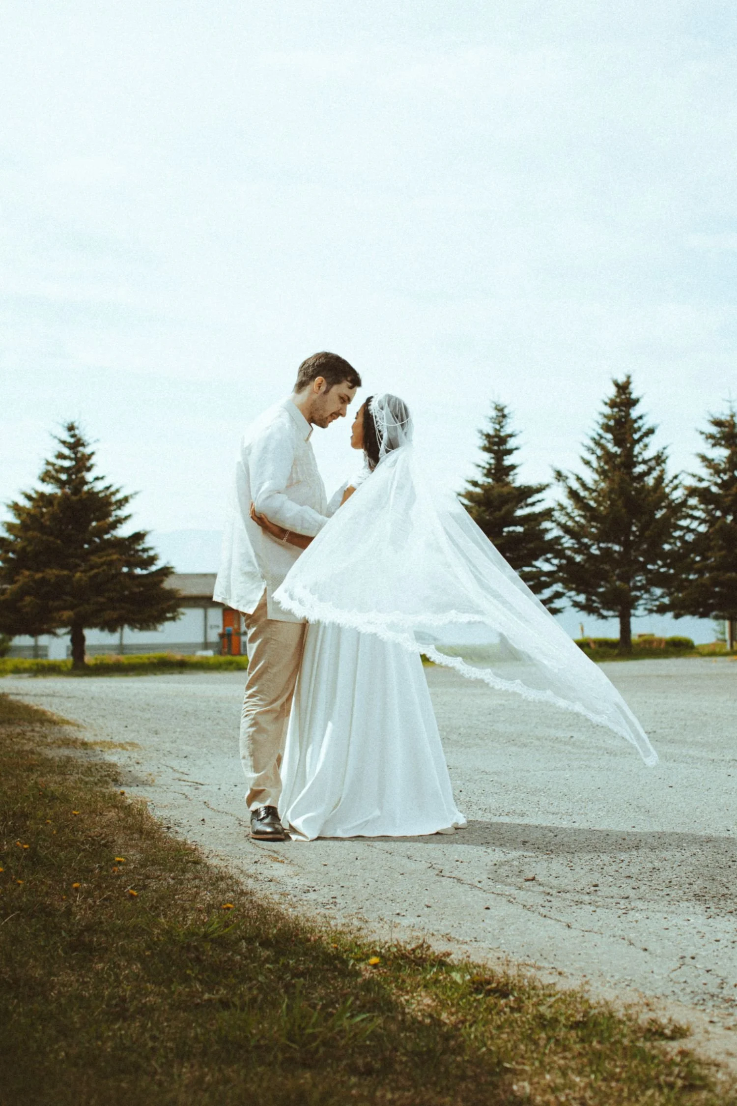 A bride and groom are standing close together outdoors, looking into each other's eyes, with the bride wearing a white wedding dress and veil, and the groom in a white shirt and beige pants, surrounded by pine trees.