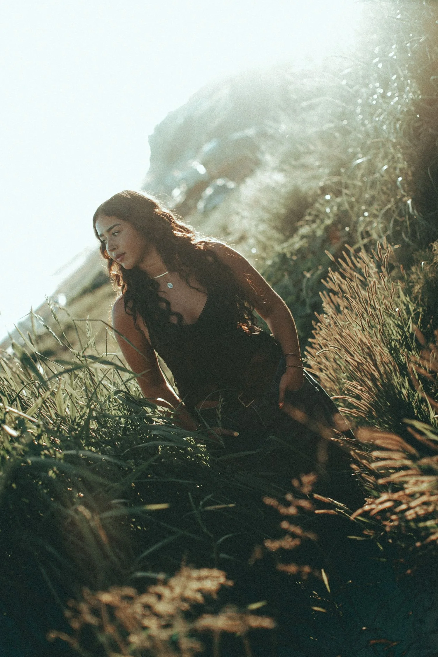 A young woman with long, wavy hair wearing a black top and jewelry, sitting in a field of tall grass with sunlight illuminating the scene from behind.