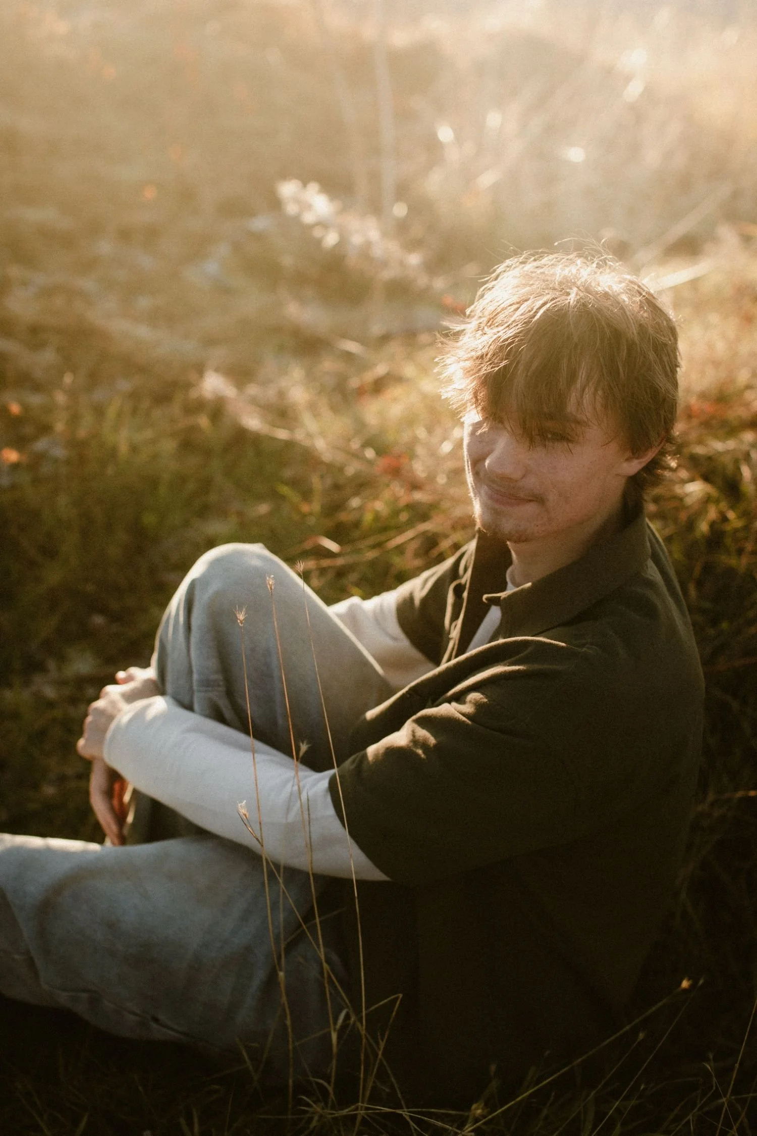A young man with light brown hair and a slight smile sitting outdoors in a field during sunset, wearing a black and white jacket and white pants, surrounded by tall grass.