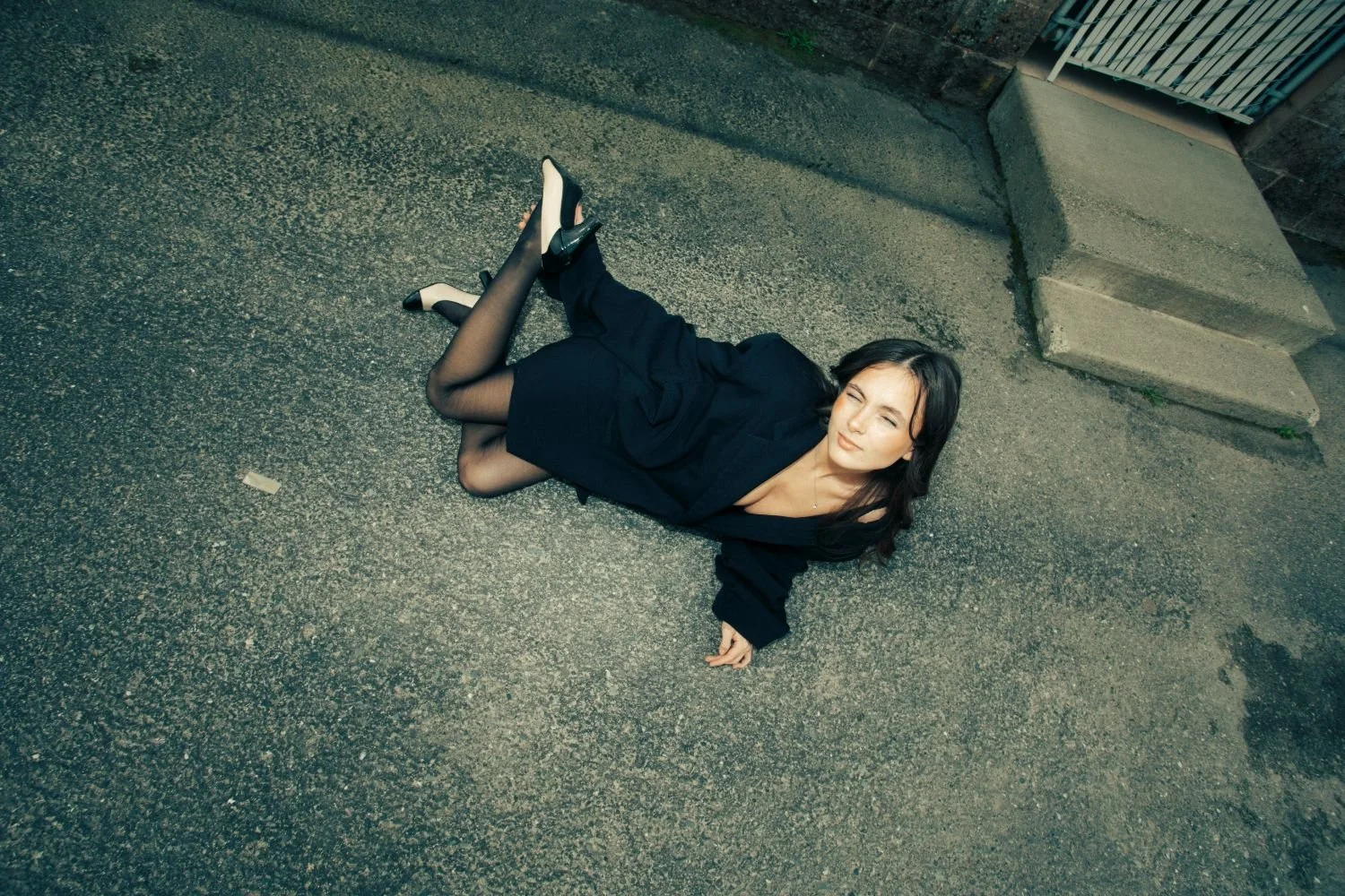 A woman lying on the ground outdoors, wearing a black dress, black stockings, and black shoes, with her hair loose, looking up and squinting at the camera.