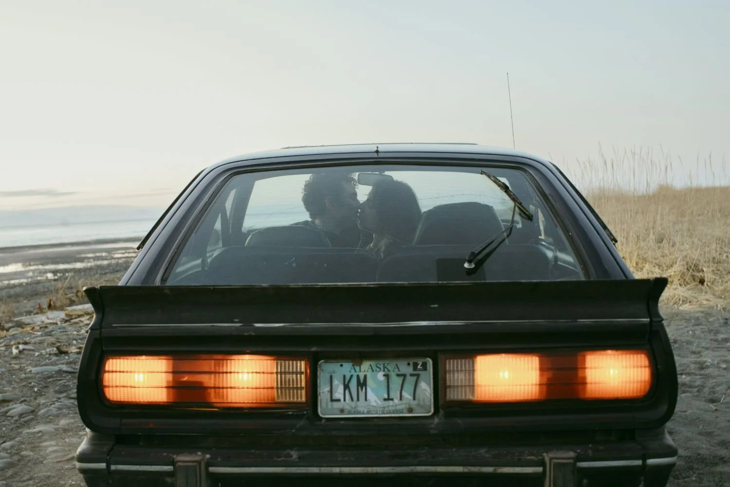 Back of a black vintage car with two people inside, close together, with a love or romantic expression, on a beach or coastal area with dry grass and ocean in the background, Alaska license plate LKM 177.