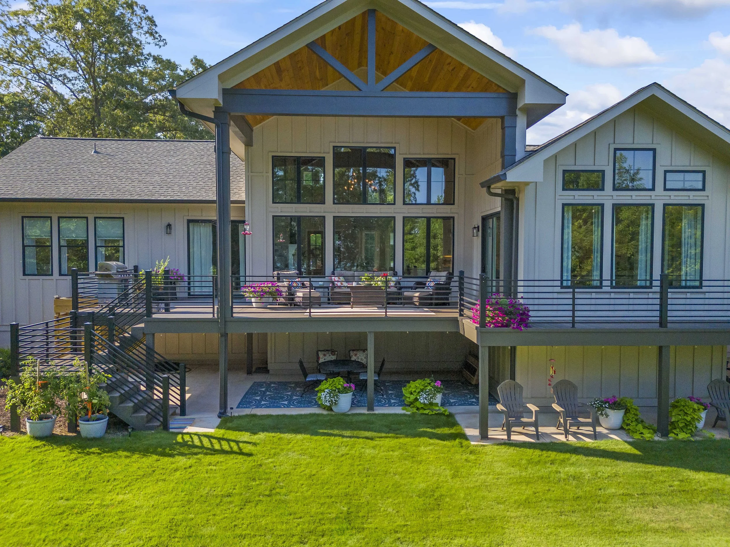 Backyard view of a modern house with large windows, wooden accents, outdoor patio furniture, flower pots, and green lawn.
