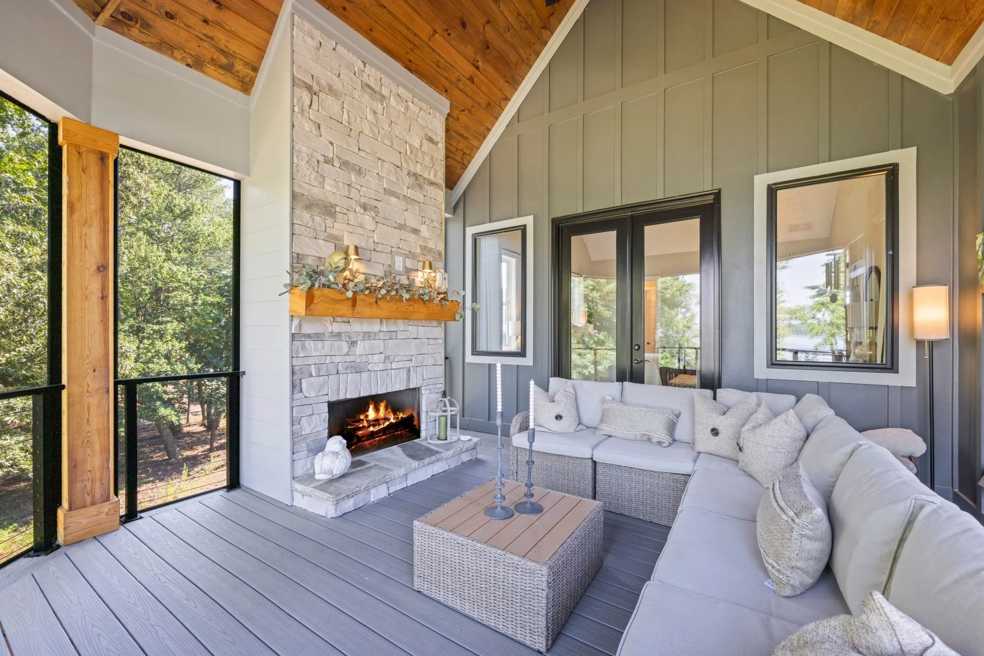 A cozy outdoor patio with a stone fireplace, white couch with pillows, a wicker coffee table, and large windows showing green trees outside. The ceiling and wall are made of wood and painted gray.