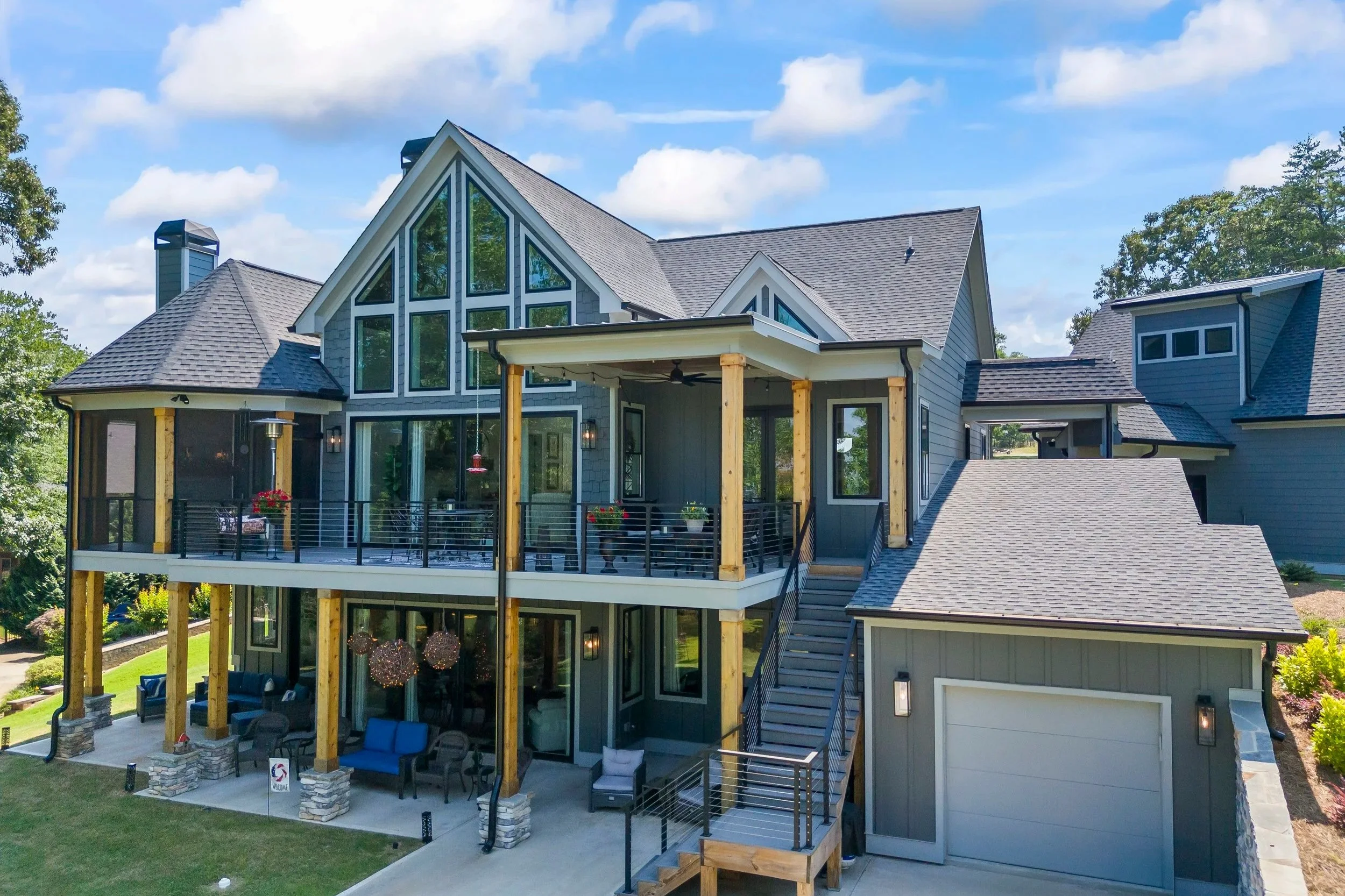 A modern multi-story house with large windows, a spacious deck, and outdoor seating area. The house has a gray exterior with a steep roof and wooden support beams on the deck. The backyard features a lawn, trees, and landscaped garden.