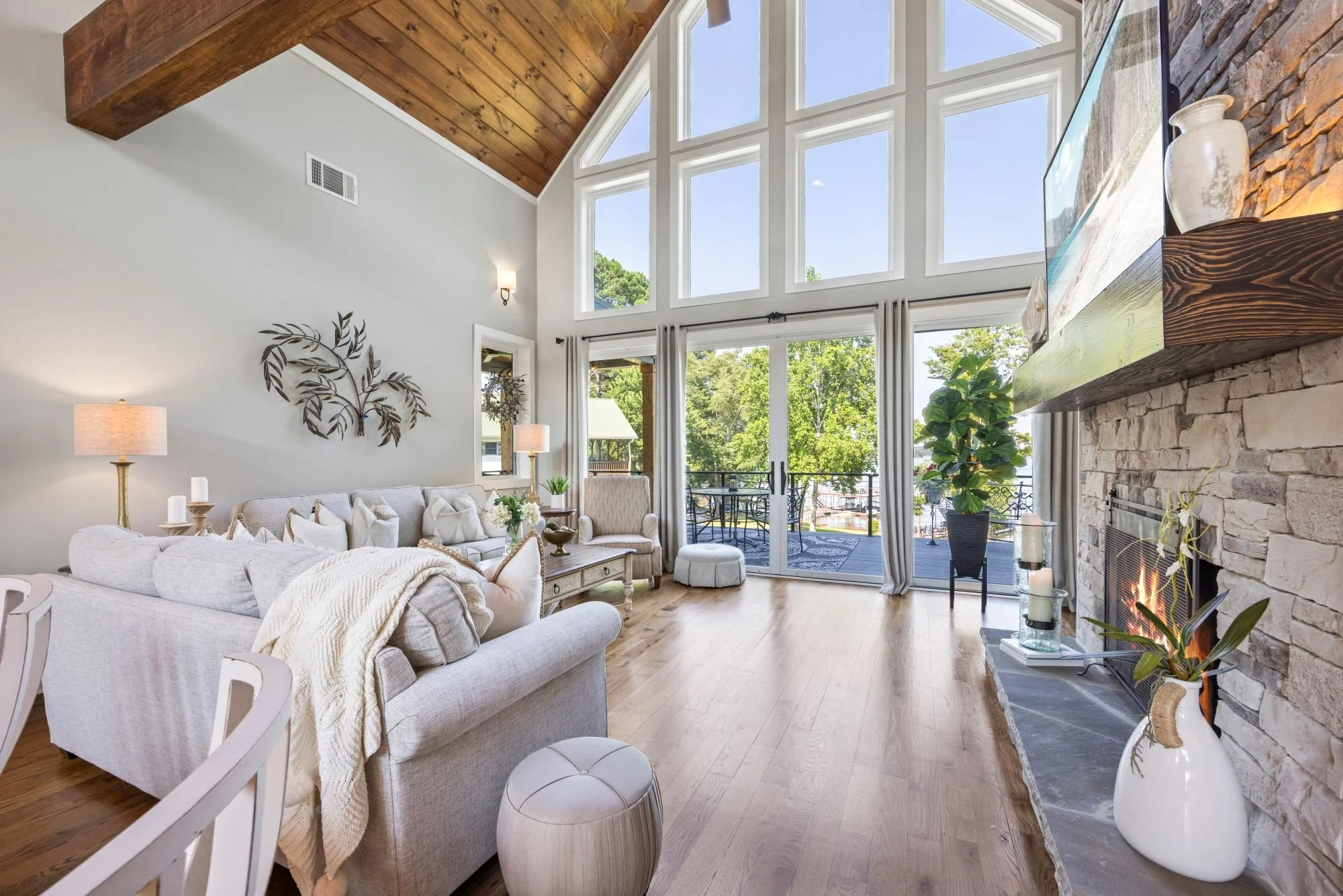 Living room with a high vaulted wooden ceiling, large geometric windows, a stone fireplace with a fire burning, a beige sofa with pillows, a white upholstered ottoman, potted plants, and decor items including lamps and vases.