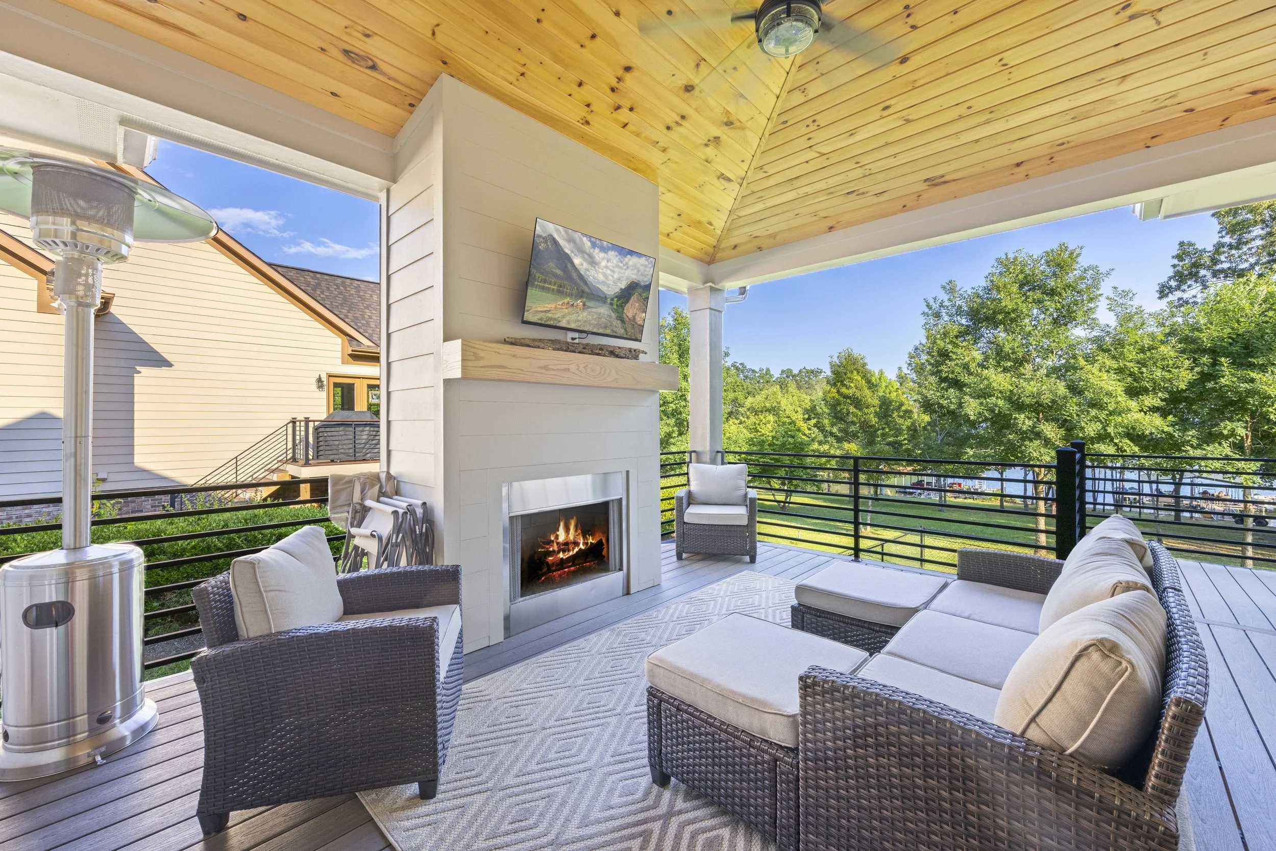 Outdoor patio with wicker sofas and chairs, a fireplace, and a mounted flat-screen TV on a white wall. The ceiling is wooden, and the patio is surrounded by green trees and a railing.