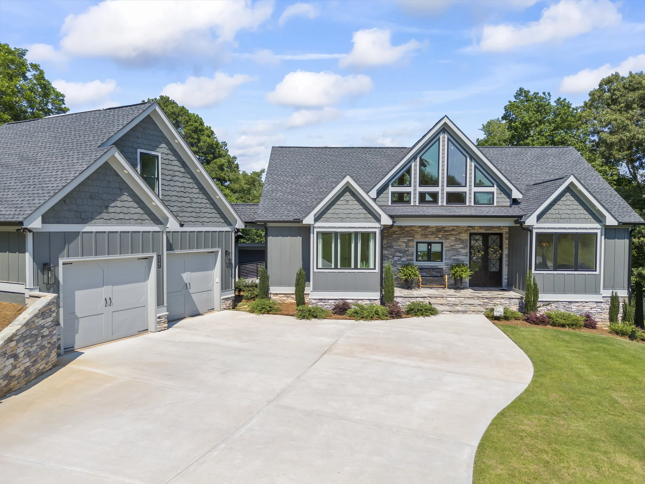Front view of a modern house with a large driveway, gray siding, and a stone accent wall, surrounded by green lawns and trees, under a partly cloudy sky.
