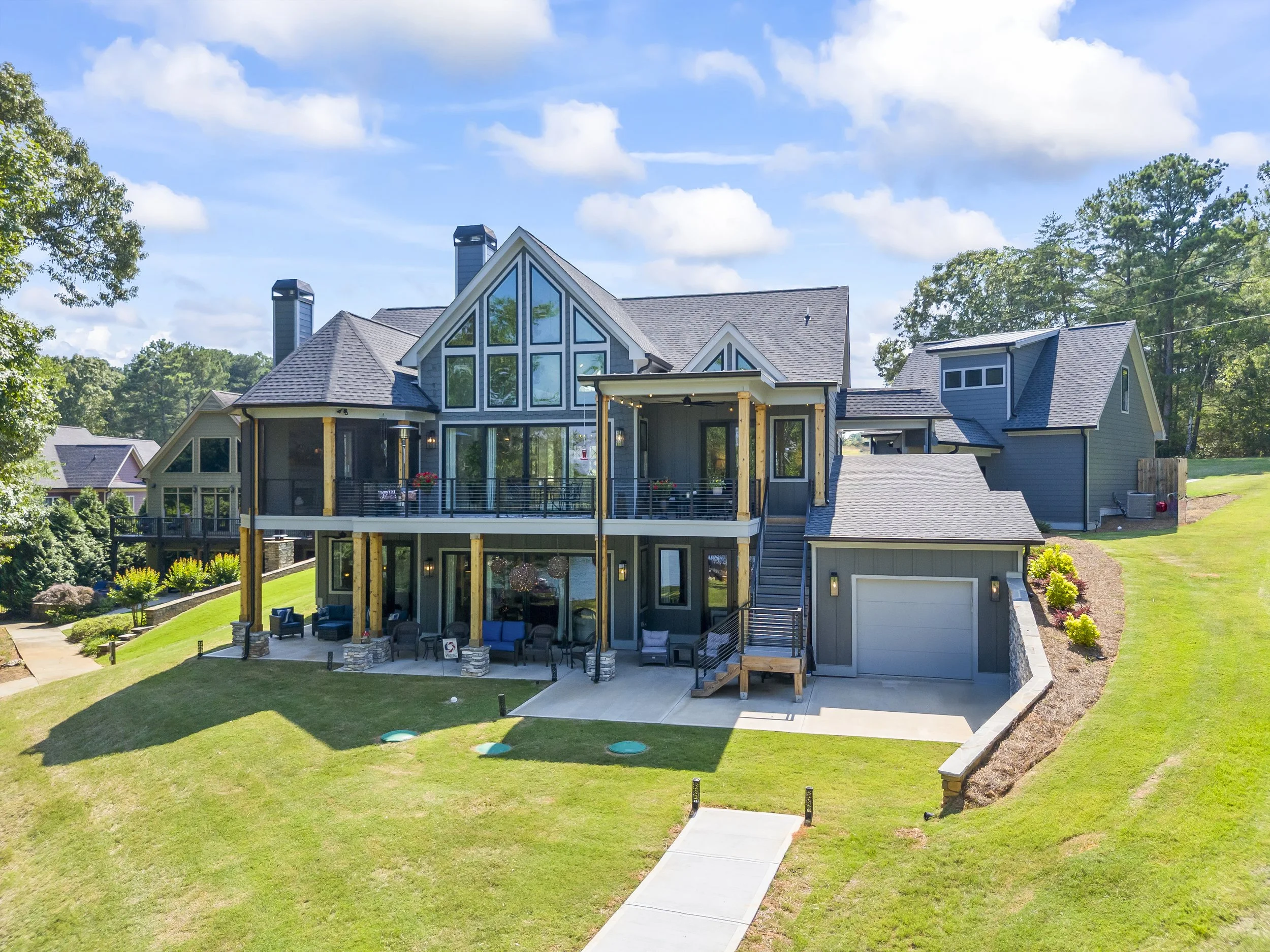 Modern house with large glass windows, surrounded by a well-maintained green lawn and trees, with a partly cloudy blue sky overhead.