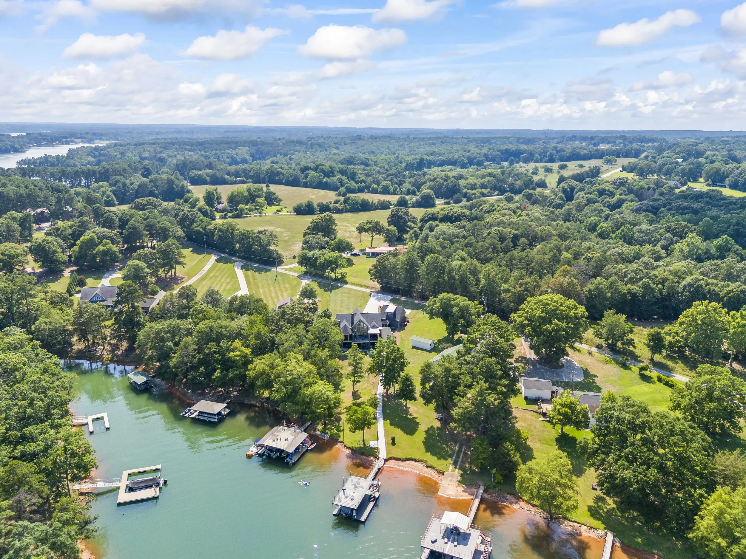 Aerial view of a lush green landscape with trees, houses, and a river with boat docks, under a partly cloudy sky.