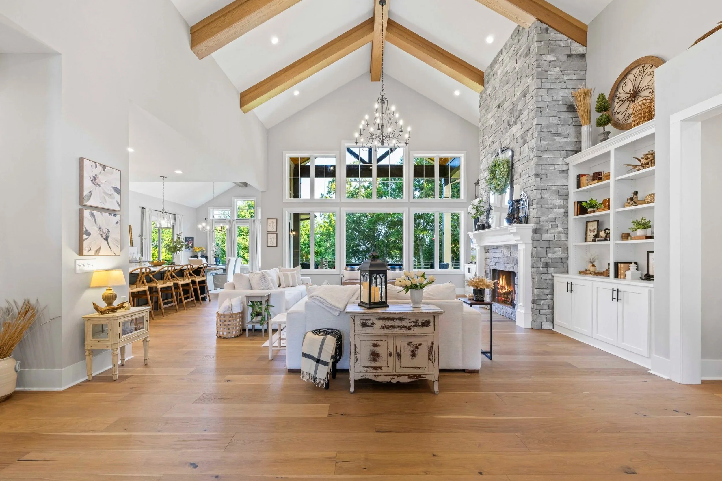 Bright living room with high vaulted ceiling, exposed wooden beams, large windows, and a stone fireplace. Decor includes white furniture, wall art, plants, and a chandelier.