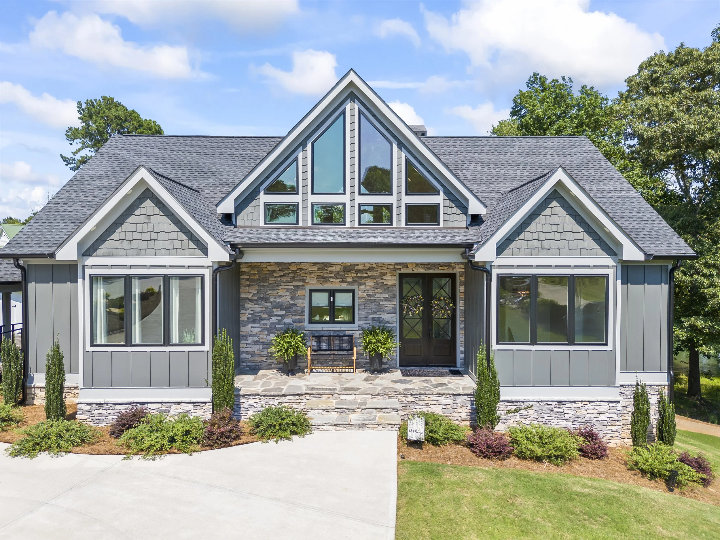 Front view of a modern house with a peaked roof, large dormer windows, gray siding, stone accents, and a small porch with plants and a bench.