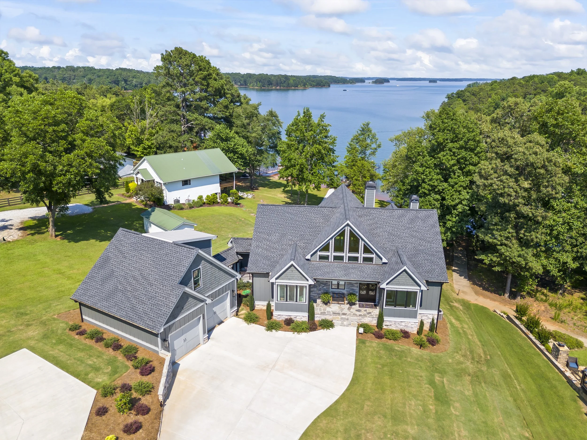 Aerial view of a lakeside residential neighborhood with modern gray houses, manicured lawns, trees, and a large body of water in the background under a partly cloudy sky.