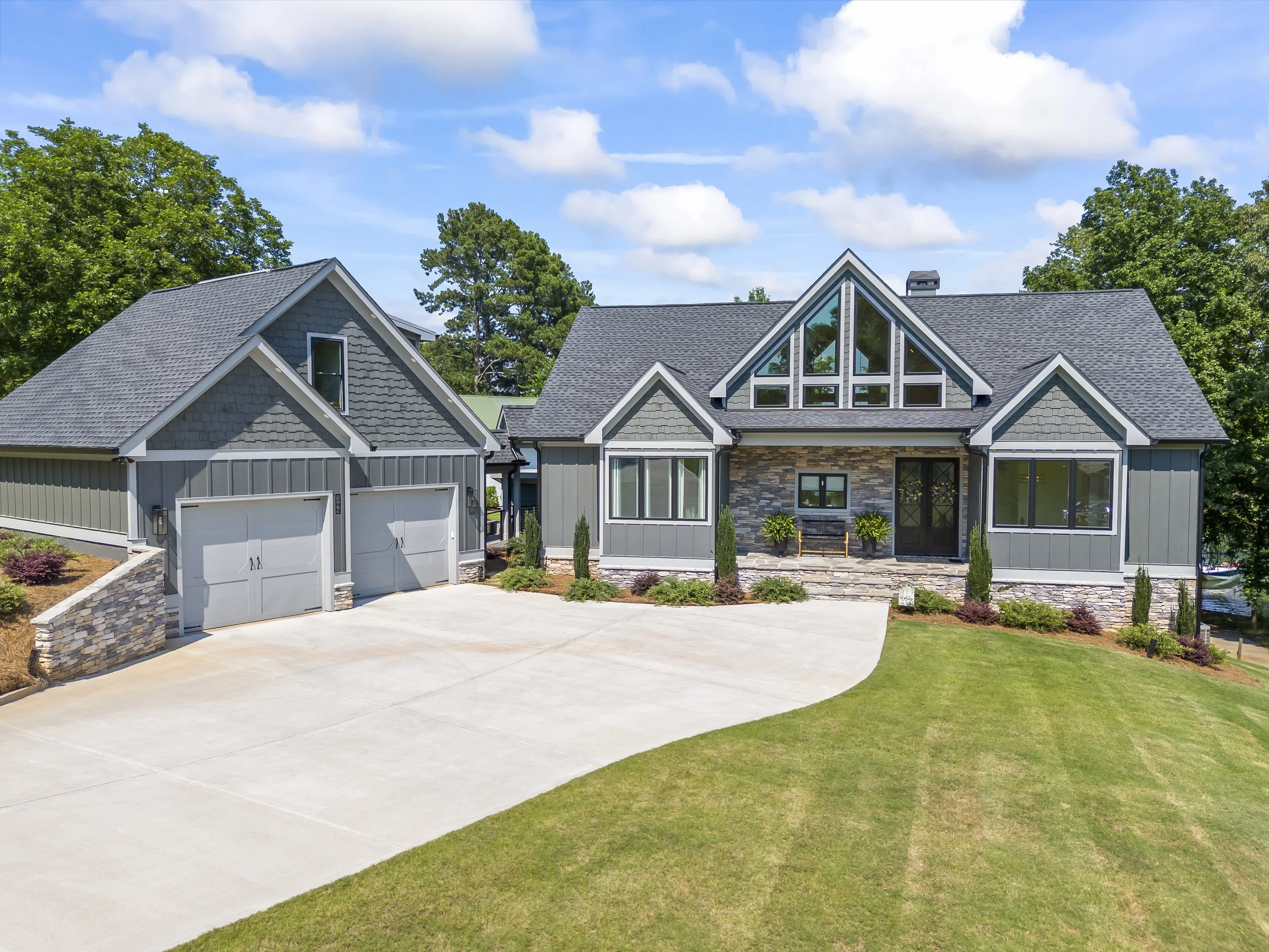 A modern house with gray siding, a stone accent wall, large windows, and a steeply pitched roof, surrounded by green grass and trees under a partly cloudy sky.