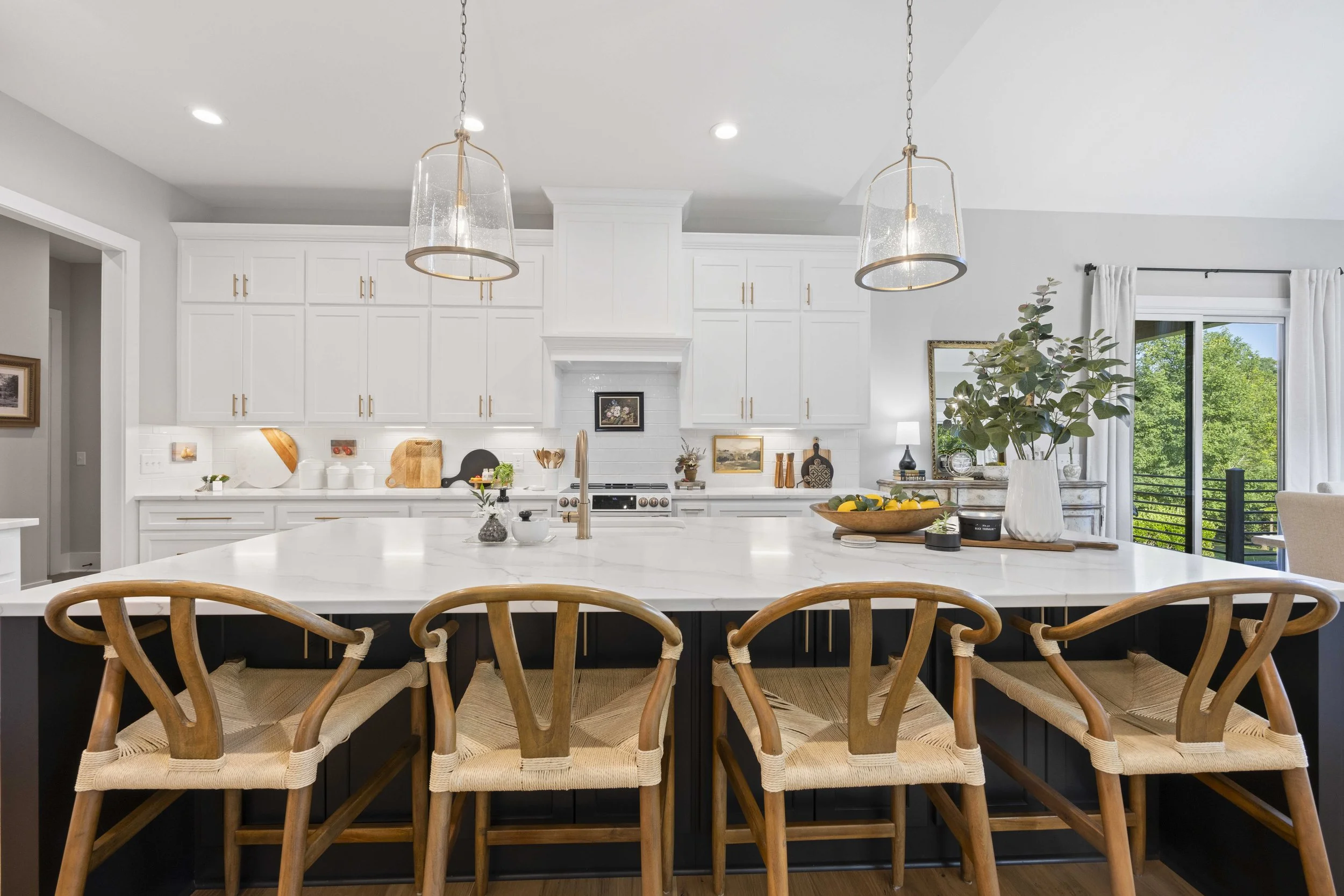 Modern kitchen with white cabinets, a large island with a white marble countertop, and wooden chairs. Two pendant lights hang above the island. There's a window with white curtains and outdoor trees visible.