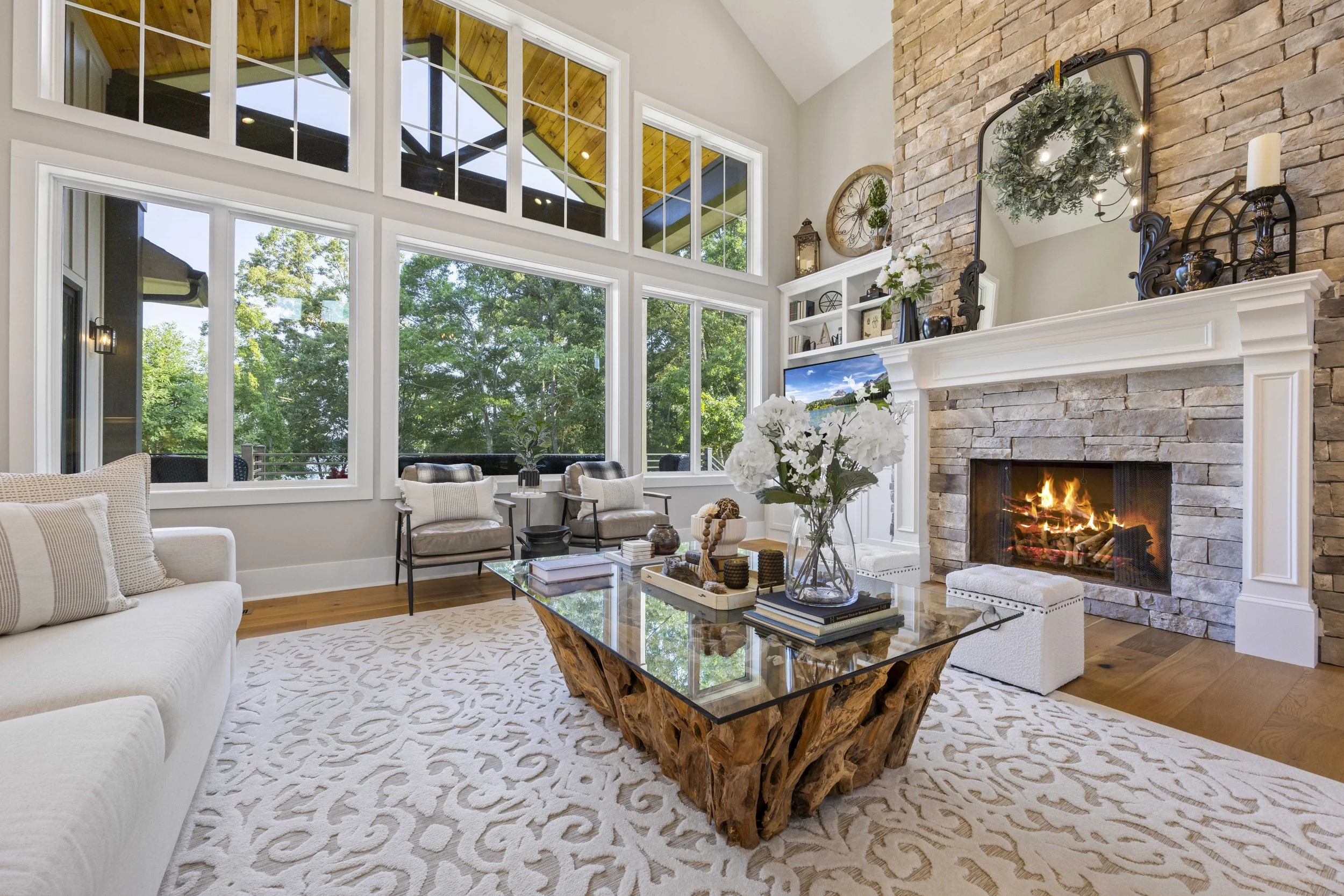Living room with large windows, a stone fireplace with a wreath, a white sofa, a coffee table with decorative items, and lush green trees visible outside.
